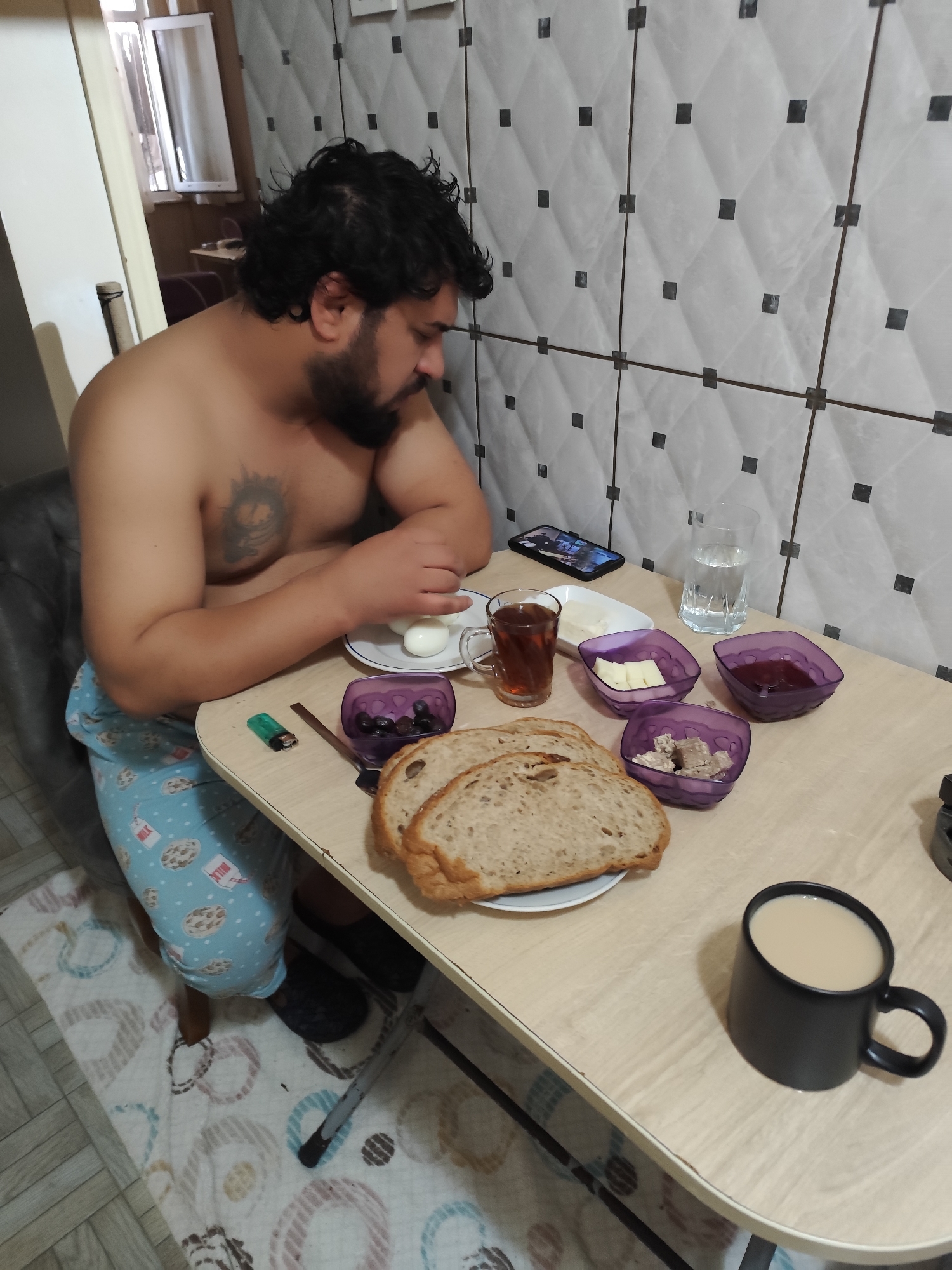 A man with dark hair and a beard sits at a table. He is shirtless and wearing patterned pajamas. On the table are two slices of bread, a plate of boiled eggs, a glass of water, tea, and several small bowls containing olives, butter, jam, and halva. There is also a black mug of tea and a lighter on the table. A cell phone is lying on the table. The location is likely a residential building in Sarıyer, Turkey, judging by the tile pattern on the wall and the presence of Turkish breakfast items.