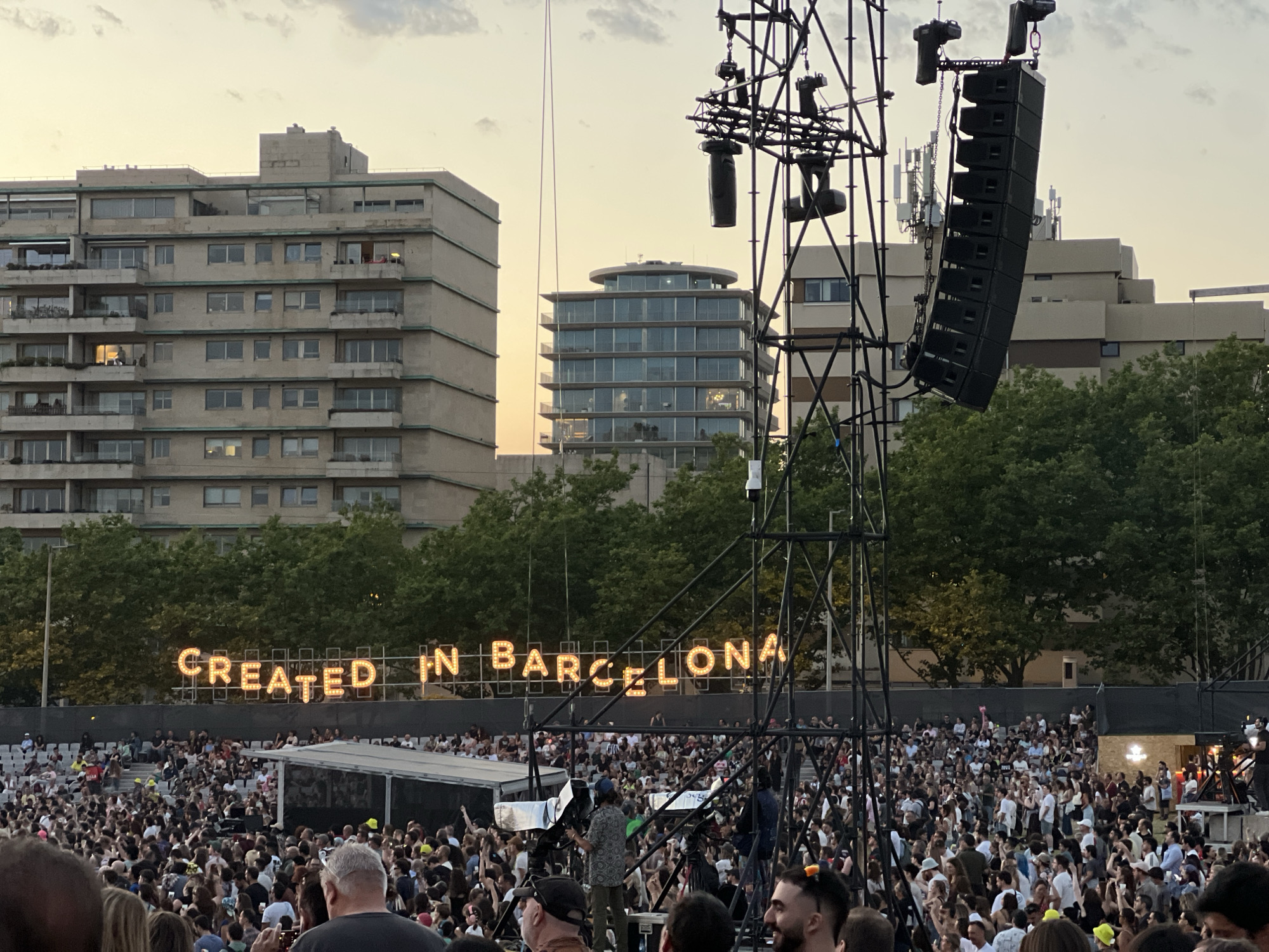 The image captures an outdoor concert scene. In the foreground, a large crowd of people are visible, many of them facing towards a stage area which is not fully visible. Above the crowd, a large metal structure with speakers and lighting equipment is prominent. In the background, there are several modern buildings and trees. A large sign with illuminated letters spelling out "CREATED IN BARCELONA" is visible across the scene, suggesting a connection to the city of Barcelona. The sky is partly cloudy with a soft, warm light, indicating it is likely late afternoon or early evening. The image does not depict the city of Porto, Portugal.