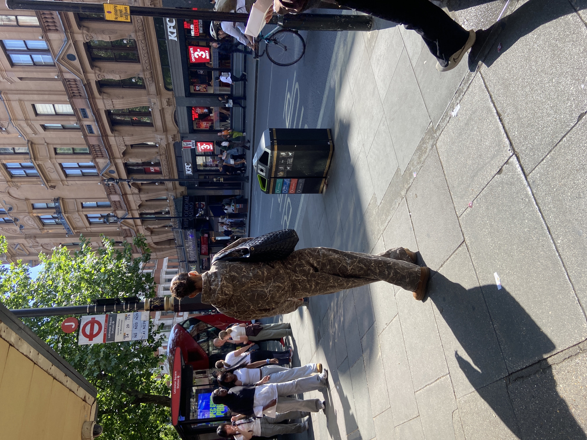 The image shows a street scene in Leicester Square, London, United Kingdom. People are standing at a bus stop with a sign clearly indicating "Leicester Square Station." A red double-decker bus is visible behind the group of people at the bus stop. A person in a patterned brown outfit is walking on the sidewalk. Across the street, there is a KFC restaurant with a sign indicating "OPEN UNTIL 3 AM DAILY." A black trash bin stands on the sidewalk between the KFC and the bus stop. The building behind the KFC has a classic architectural style with detailed window frames and a brown facade.
