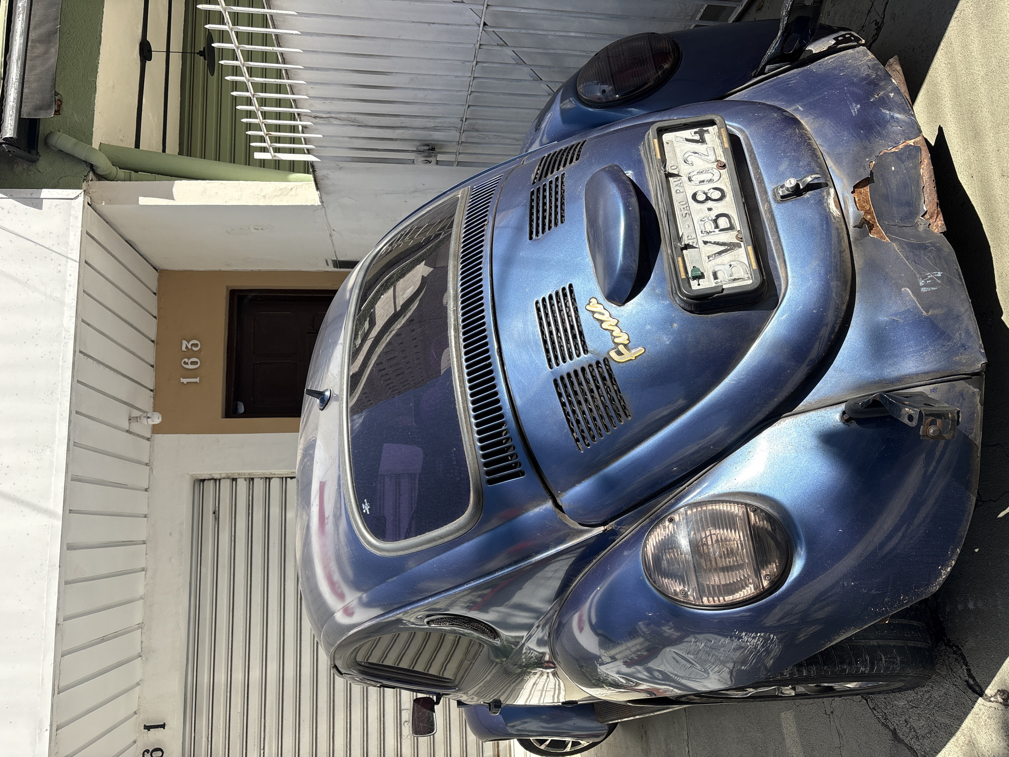 A blue Volkswagen Beetle, possibly a Fusca, is parked in front of a residential building. The building has a white facade with numbers "61" and "163" visible above garage doors. The Beetle's rear is facing the camera, and it shows signs of wear and tear, with rust visible on the bumper. A license plate is mounted on the rear, displaying "BVB-8024" and "SAO PAULO". The scene appears to be in an urban environment.