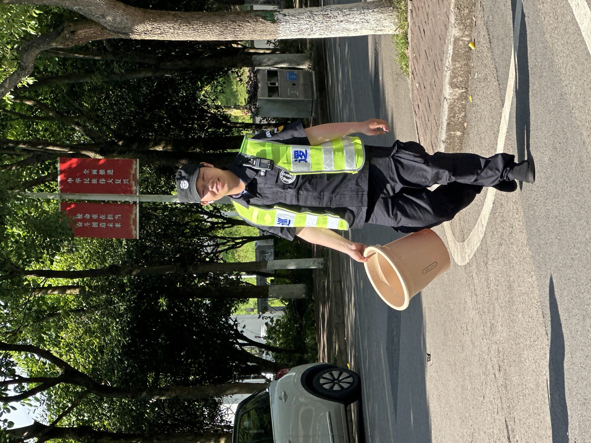 A young man in a dark blue uniform, a fluorescent yellow reflective vest, and a dark cap stands outdoors on a sunny day in Jiangjin, China. He is smiling towards the camera while holding a light brown plastic bucket in his right hand. A walkie-talkie is clipped to his vest, which features the Chinese characters "城管" (chéngguǎn), indicating urban management.
The man is positioned on a paved road or pathway, with a brick sidewalk to his left. Behind him and to his left, a white car is partially visible. Lush green trees line the background and the left side of the scene. To his left, behind him, a prominent red sign with white Chinese characters is visible. The top text reads: "全面推进 中华民族伟大复兴" (Comprehensively advance the great rejuvenation of the Chinese nation). The bottom text reads: "牢记使命 勇于担当 争创一流 赶超进位" (Keep the mission firmly in mind, be brave in undertaking responsibilities, strive to create first-class achievements, catch up and overtake). A grey municipal trash can is also visible in the background.
The bright sunlight casts distinct shadows on the ground, suggesting it is mid-morning or afternoon on a clear day. The word "JIA YU" is faintly visible on the side of the bucket he is holding.