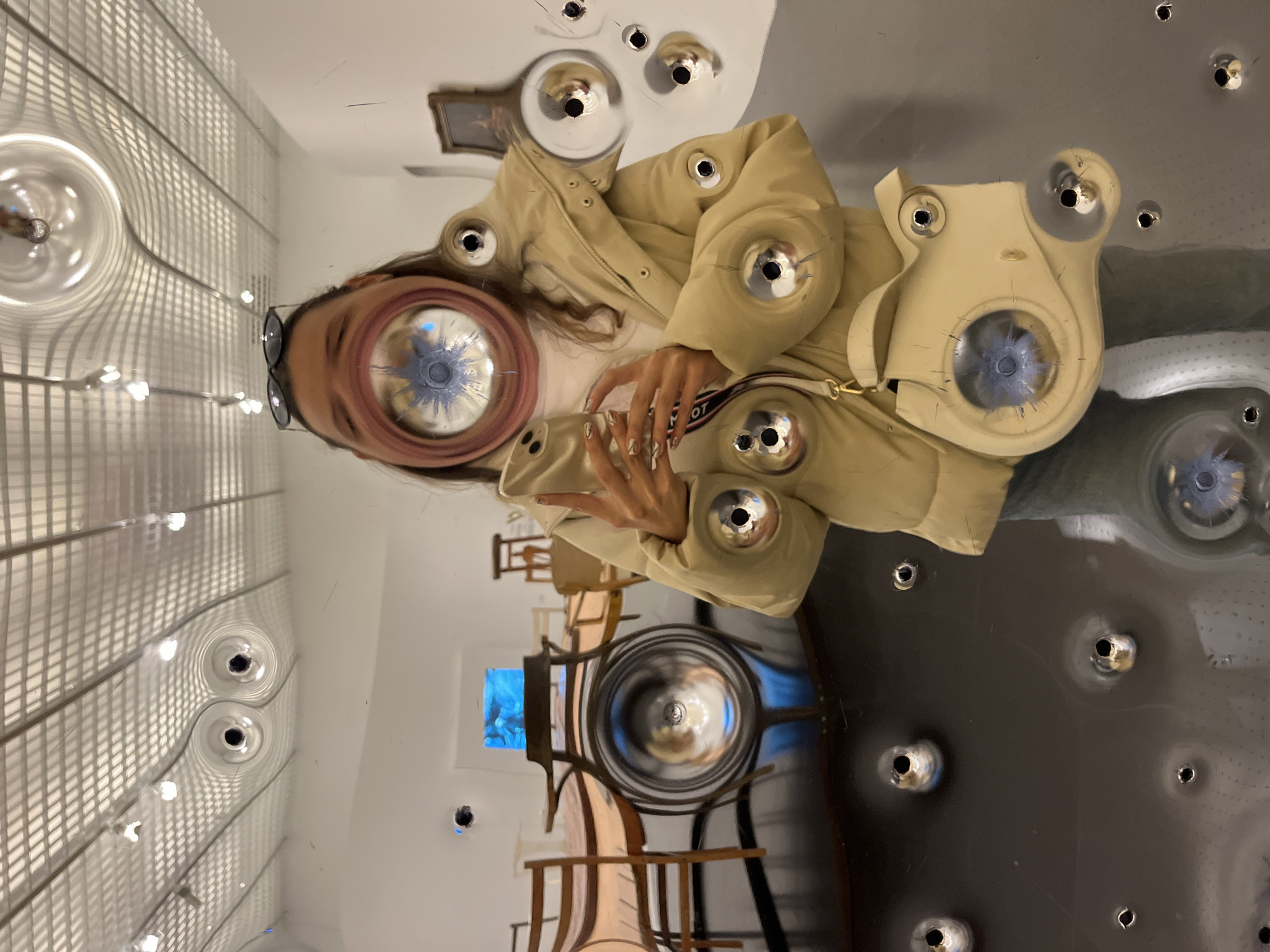 The image captures a warped reflection in a shiny surface, possibly stainless steel, in what appears to be a room at the Centre Pompidou-Metz in Metz, France. The reflection shows a woman wearing glasses and a beige jacket with numerous circular metallic details that seem to be reflecting the light fixtures on the ceiling. The surroundings, also reflected, include a hallway with rows of spherical light fixtures and some furniture. The shiny surface is pockmarked with small circular holes, distorting the reflected image and creating surreal effects.