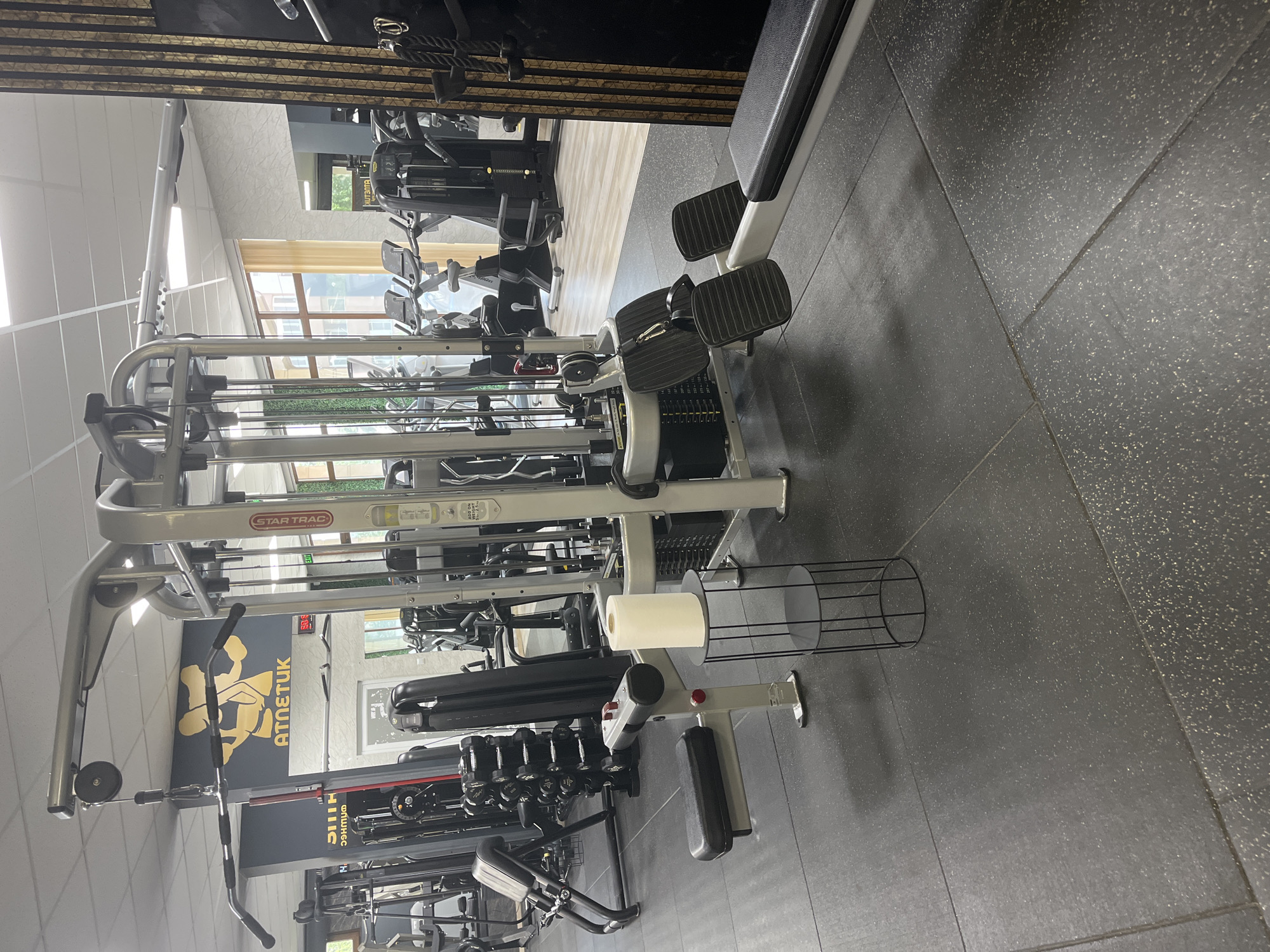 The media file displays the interior of a well-equipped gym, devoid of any people. The primary subjects are various pieces of fitness equipment, the gym floor, and parts of the walls and ceiling.
In the foreground and central area, a large multi-station cable machine, branded "STAR TRAC," dominates the view. It features several weight stacks, pulleys, and attachments, including a lat pulldown bar and footplates that appear to be part of a seated row station. Next to this, a white paper towel roll is seen on a black wire dispenser, suggesting a focus on hygiene. To the left, a rack of dumbbells of varying weights is visible. The floor throughout the gym is covered with dark grey rubberized matting, speckled with lighter flecks, characteristic of a professional fitness environment.
Further back, more gym equipment can be seen, including benches and other strength training machines, some with black upholstery. Natural light streams in through large windows on the far wall, illuminating the space and suggesting it is daytime. Outside the windows, some greenery and building structures are faintly visible.
A prominent gold-colored logo is affixed to a dark wall, depicting a stylized figure engaged in weightlifting, with text beneath it that reads "ATMETUK" (possibly "ATLETUK," meaning 'athlete' in some languages). The ceiling is light-colored with grid patterns, and some sections of the wall feature decorative wooden slats. Another small, possibly partially obscured text, "AIR ON," can be seen on one of the machines. The overall impression is a modern and functional fitness facility.
