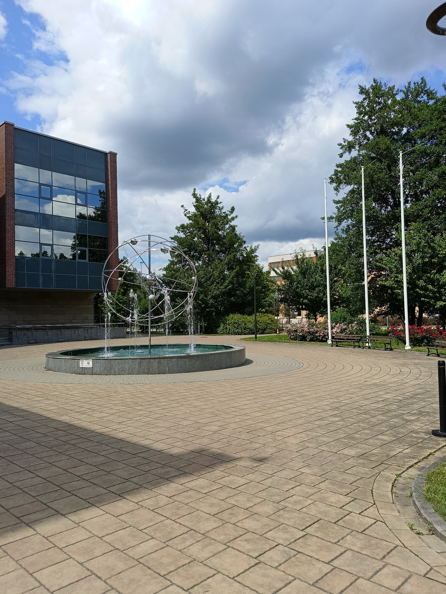 A contemporary building with a red brick facade and large dark-tinted windows stands to the left of a circular fountain. The fountain features a metallic, spherical sculpture with multiple rings and orbiting spheres, from which water is spraying upwards and outwards. The fountain is set in a paved courtyard with interlocking beige rectangular stones arranged in concentric circles and straight lines. Several mature green trees are visible in the background, as well as a large, multi-story building with lighter colored walls and windows. Three tall, white flagpoles stand in the middle ground. Two park benches are situated in front of a bed of pink and red flowers. The sky is partly cloudy with patches of blue visible. The scene appears to be a public space or campus, possibly located in Krakow, Poland.