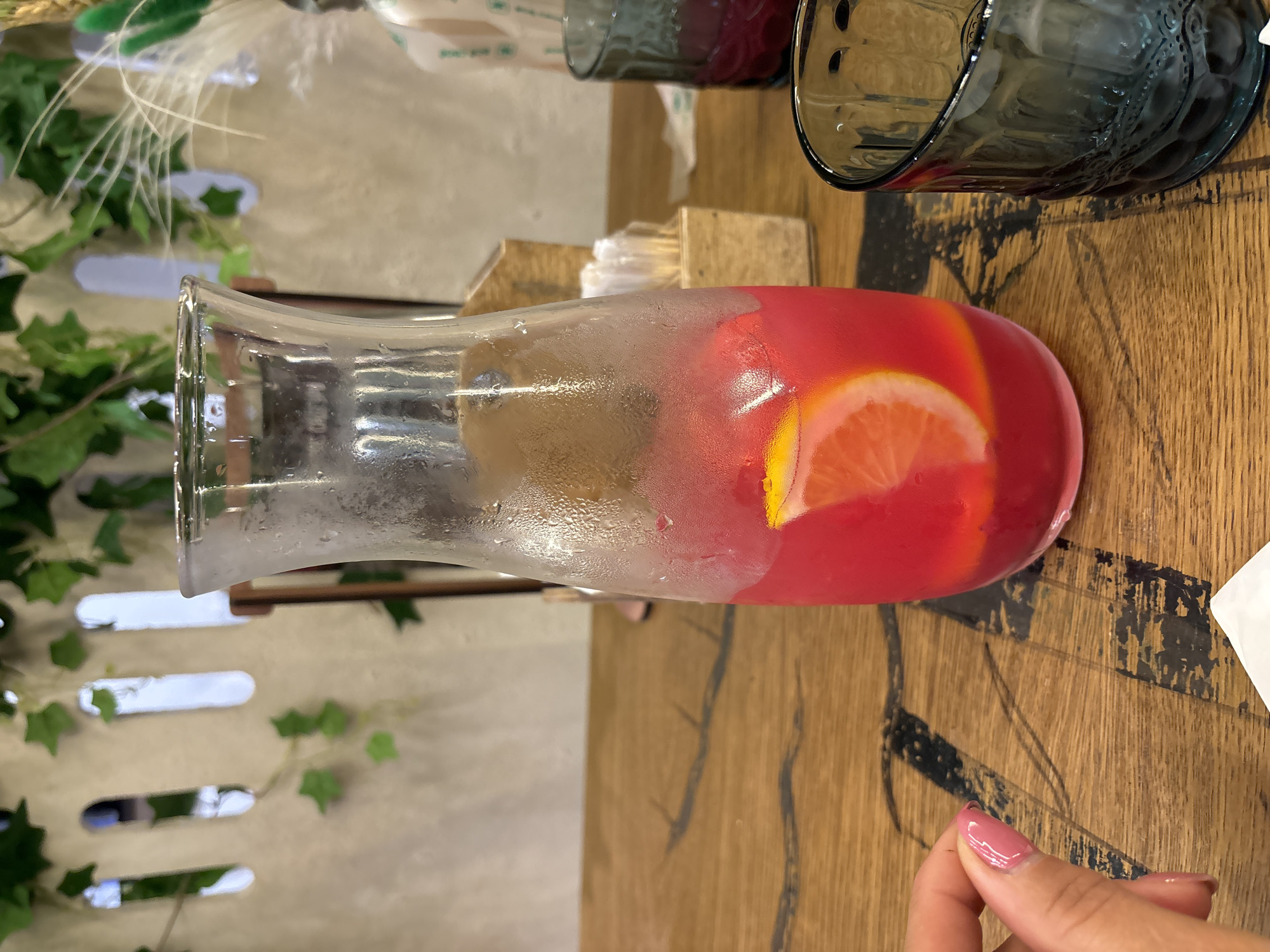 The image shows a close-up of a table at a restaurant. In the foreground, a hand with pink nail polish is visible on the left side of the frame. The main subject is a clear glass carafe filled with a pink beverage, containing slices of citrus fruit. The carafe is covered in condensation. To the right of the carafe is a dark blue glass, also with condensation. The table surface is made of wood with a dark, abstract pattern. In the background, a light-colored wall with decorative cutouts and artificial ivy is visible. A wooden holder with white straws is positioned behind the carafe. The scene suggests an indoor dining setting. There is no information in the image to indicate it is located in Izhevsk, Russia.