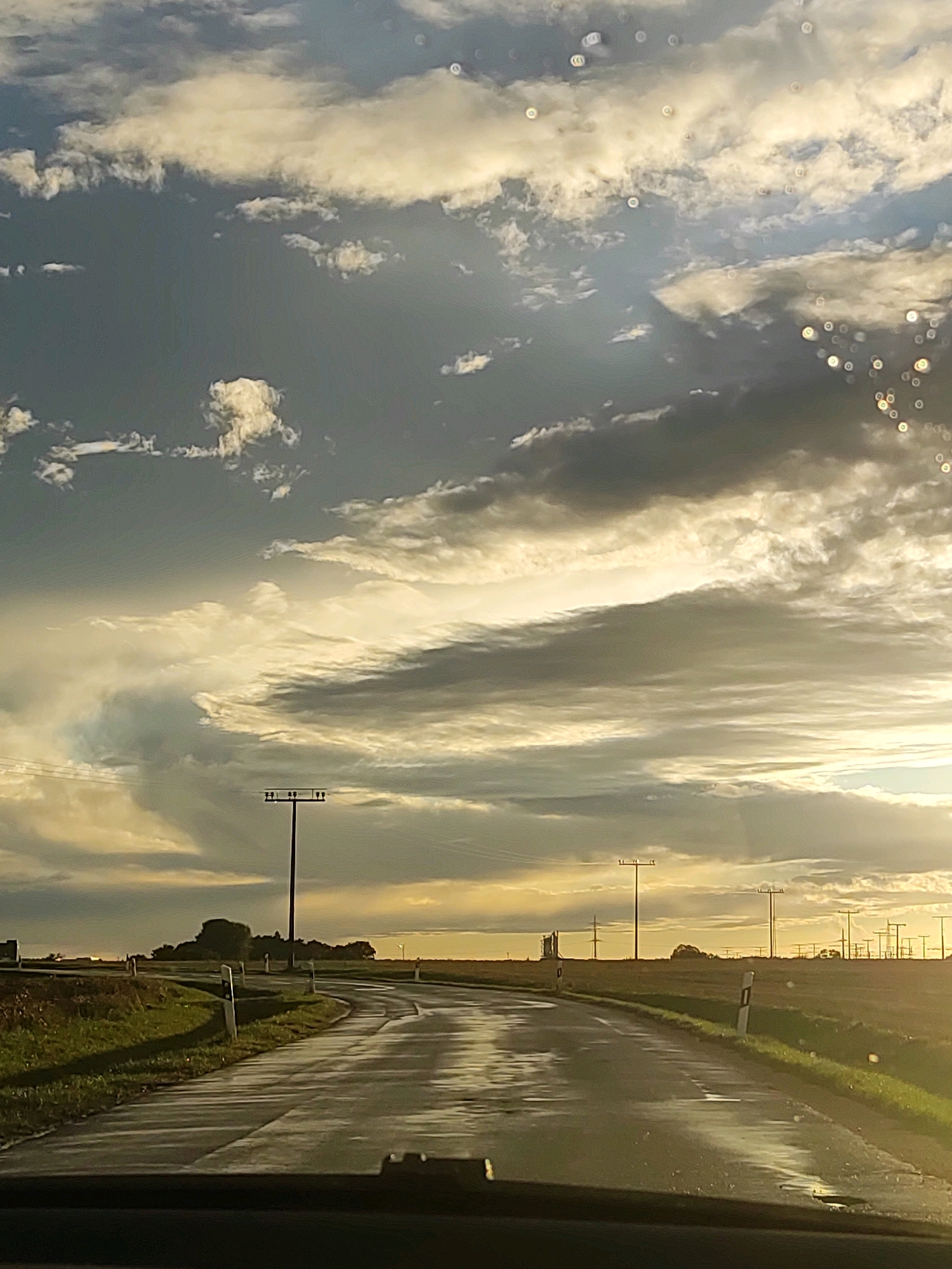 The image depicts a rural road curving through a landscape with a dramatic sky. The road appears wet, suggesting recent rain. The sky is filled with dynamic clouds, illuminated by what seems to be the setting or rising sun, casting warm light and shadows. Electrical poles with multiple wires are visible along the side of the road and in the distance, indicating a rural or semi-rural setting. The presence of roadside markers suggests this is a paved road. The foreground shows the dashboard of a vehicle, implying the photo was taken from inside a car. There are no people or animals visible in the frame. This scene could be located on a road outside the city of Ansbach, Germany, characterized by open fields, agricultural land, and the typical infrastructure of such regions, including power lines and road signage. The overall mood is atmospheric, with the interplay of light and clouds creating a sense of a transient moment.