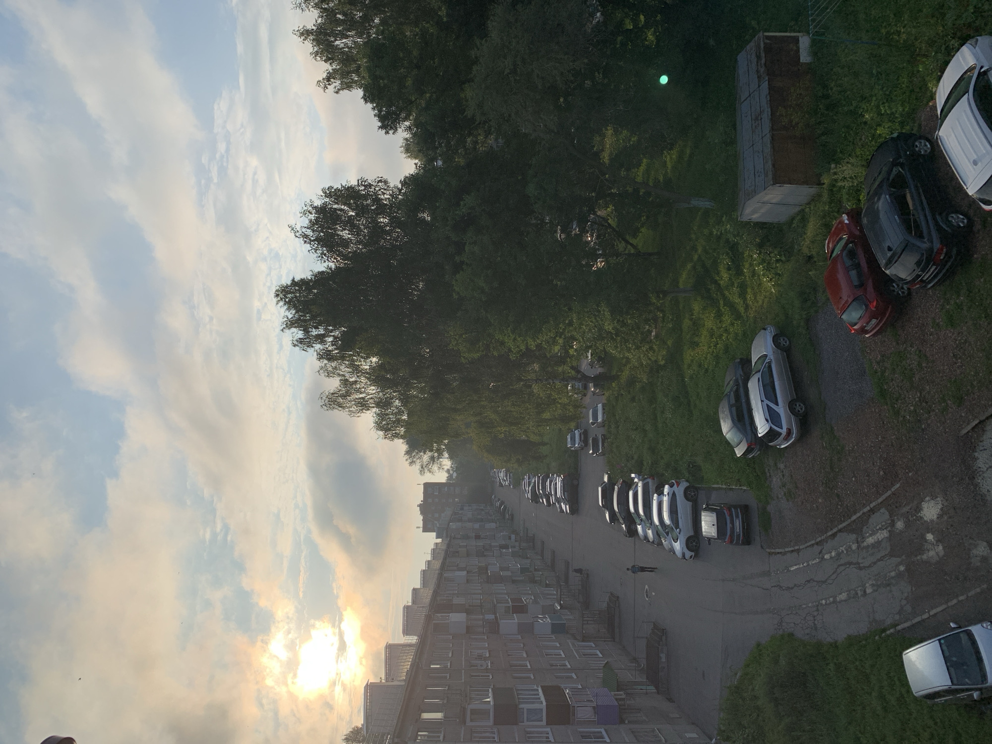An outdoor, slightly elevated, wide shot shows a residential street scene in Prokopyevsk, Russia, under a cloudy sky with the sun peeking through. The image is dominated by a long, multi-story apartment building on the left, with numerous balconies and windows. A row of parked cars lines the street in front of the building and extends into the distance. The street is paved but appears weathered and cracked in places, with patches of overgrown grass and weeds along the edges.
A single man is walking down the street away from the camera. Further down the street, a few more apartment buildings are visible. To the right of the street and cars, a line of mature trees forms a dense green backdrop. In the foreground on the right, there is a small, shed-like structure with a corrugated roof. Several cars are parked on the grass and dirt area next to the shed. A bright green spherical object, possibly a lens flare or light reflection, is visible in the upper right portion of the image. The overall impression is of a typical urban residential area during daylight, possibly early morning or late afternoon due to the sunlight filtering through the clouds.