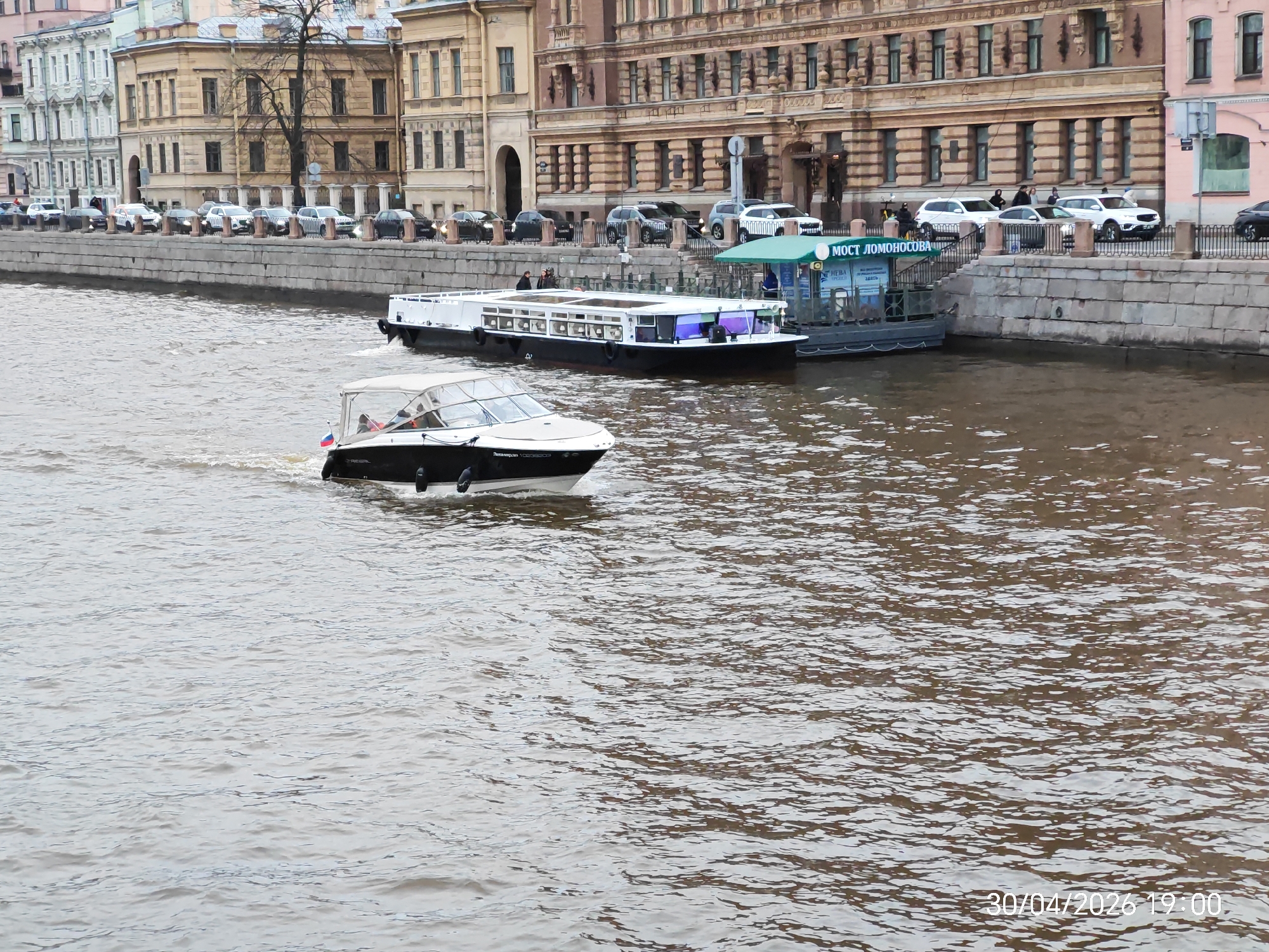 This image depicts an urban waterway scene, likely a canal or river, flanked by historical buildings and an embankment. In the foreground, a white and black motorboat with a clear canopy travels across the water from left to right, generating a visible wake. A small flag with the colors of the Russian tricolor is affixed to its front, and partially visible text on its hull includes "REGAL" and what appears to be "Портовик".
Along the right bank, a sturdy stone embankment topped with a black metal fence supports a street where several cars, mostly SUVs, are parked or driving. Beyond the street, a row of multi-story buildings showcases classical European architecture with ornate detailing, in varying shades of beige, light yellow, and soft pink. A bare tree stands in front of one of these buildings.
Closer to the right bank, two other boats are present. A large, white and black passenger boat, featuring numerous windows, appears to be either docked or slowly maneuvering. Adjacent to it, a smaller boat with a distinctive green canopy is docked. This green canopy displays Cyrillic text that reads "МОСТ ЛОМОНОСОВА," translating to "Lomonosov Bridge." Several people are visible on the decks of these boats and on the embankment.
The water is a brownish-grey hue, covered in ripples and small waves. The overall lighting suggests an overcast day, with no harsh shadows present. A digital timestamp in the bottom right corner of the image indicates the date and time as "30/04/2026 19:00," placing the scene in the early evening. The combination of architecture, visible text, and the flag suggests a location in Russia, likely St. Petersburg.