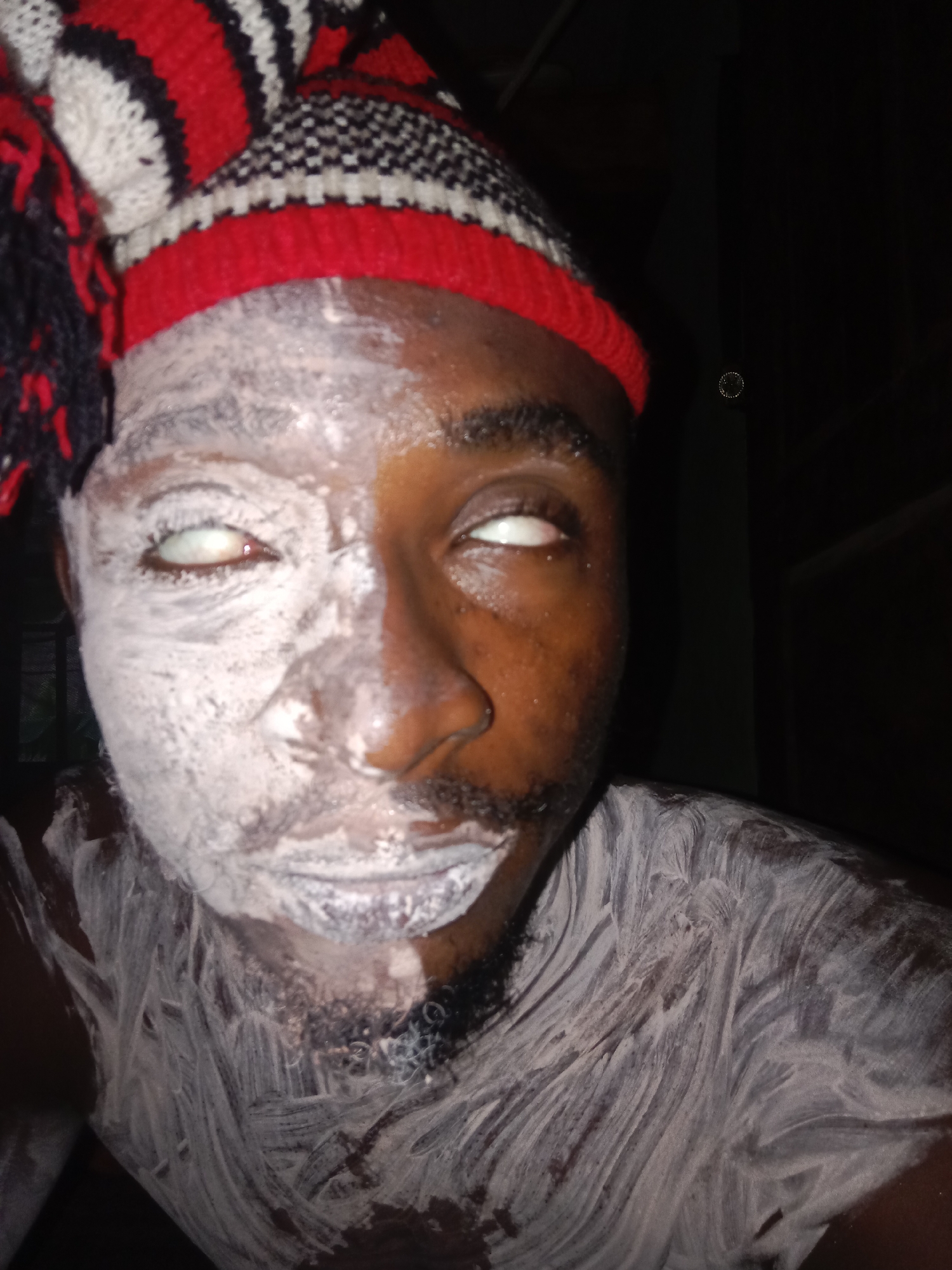 A man is seen in a close-up shot, appearing to have white cream or a white substance applied to half of his face and his upper body. He wears a red, white, and black striped knit cap. Both of his eyes are rolled back, displaying only the whites. The location is unidentified.