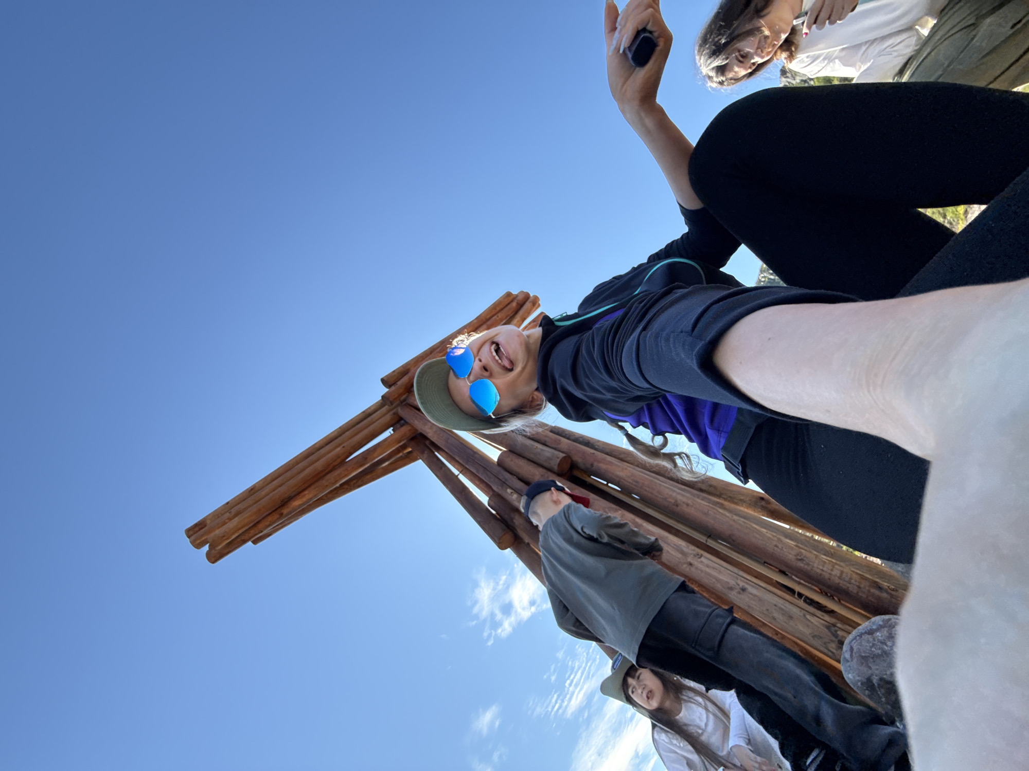 A low-angle, wide shot captures a group of four people outdoors under a clear blue sky with scattered clouds. The group is gathered around a tall, conical structure made of wooden poles. In the foreground, a woman in a black jacket, purple top, and black leggings is in the process of taking a selfie, holding a phone in her outstretched right hand. She is wearing a green cap and blue mirrored sunglasses and has her tongue sticking out playfully. To her left, slightly behind her, are two children. One child, wearing a white long-sleeved shirt and a grey baseball cap, is looking towards the wooden structure. Another child, wearing a white long-sleeved shirt and a grey hat, is also looking away from the camera. To the right of the woman in the foreground, another person, possibly a teenager or young adult, wearing a white long-sleeved shirt, is visible, looking downwards and holding something in their hand. The environment appears to be natural, with some vegetation visible in the background and a rock in the immediate foreground. The location is identified as Teriberka, Russia.