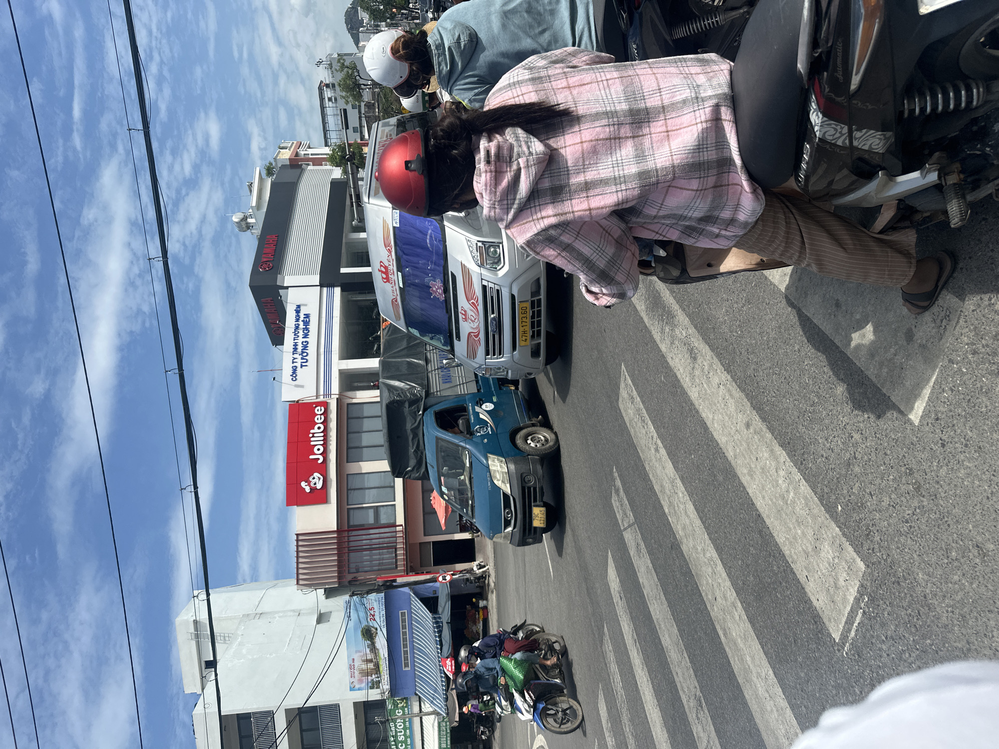 In Nha Trang, Vietnam, on a sunny day with a partly cloudy sky, a street scene unfolds at a pedestrian crossing. Two individuals are on a motorcycle in the foreground, viewed from behind. The driver wears a red helmet, and the passenger, a pink and grey plaid hoodie and striped pants. To their left, another motorcycle carries two passengers, both wearing helmets and dark clothing.
Further into the scene, a blue truck with a tarp-covered cargo bed is stopped at the intersection. To the right of the truck, a silver van with red and gold graphics on its windshield is also present. The background features commercial buildings, including a Jollibee restaurant with its distinctive red sign, and a Yamaha dealership. Overhead, black wires crisscross the sky, casting faint shadows on the asphalt road. The pedestrian crossing, marked by white stripes, dominates the lower portion of the image. The overall impression is a typical urban street scene in Vietnam, characterized by a high density of motorcycles and commercial activity.