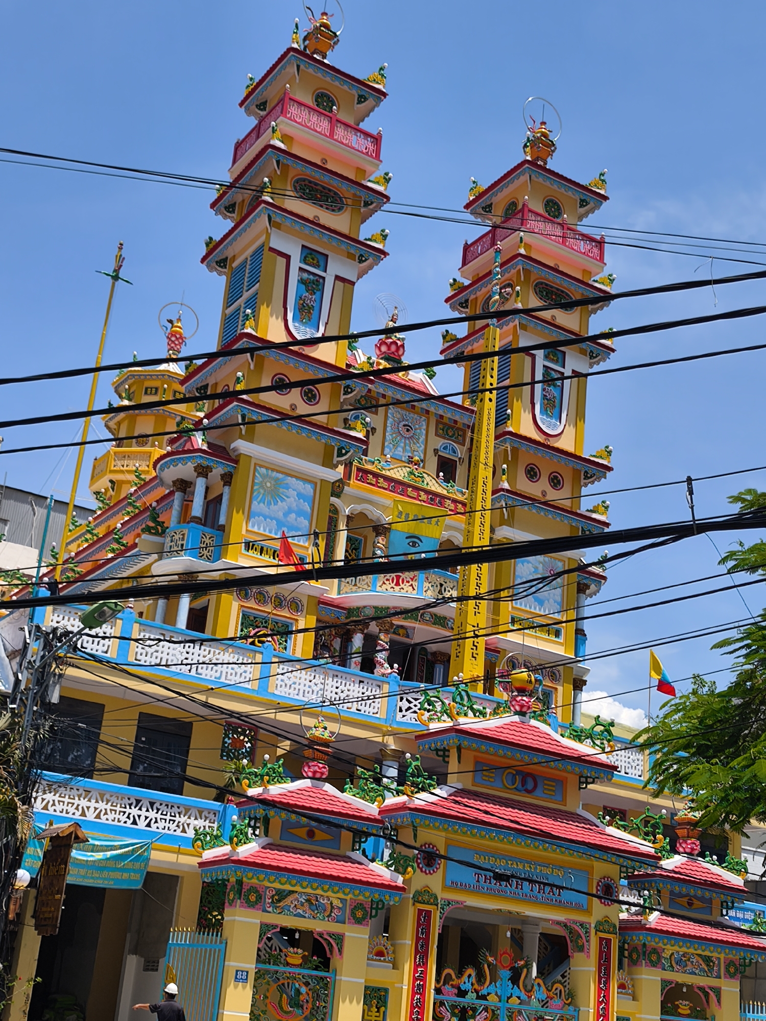 In Nha Trang Ward, Vietnam, a vibrant yellow pagoda with intricate red and blue architectural details stands prominently against a clear blue sky. The pagoda features multiple tiered roofs and towers, adorned with decorative sculptures and colorful motifs. Electrical wires crisscross the foreground, partially obscuring the view of the elaborate structure. The building exhibits ornate patterns, including depictions of celestial bodies, mythical creatures, and symbolic imagery. A small red and yellow flag is visible near the top of the pagoda, and lush green foliage is present to the right. In the lower left portion of the image, a person wearing a white hard hat and dark clothing gestures towards the pagoda. The lower section of the pagoda is decorated with a large blue banner and the number "88" is visible on a yellow pillar.