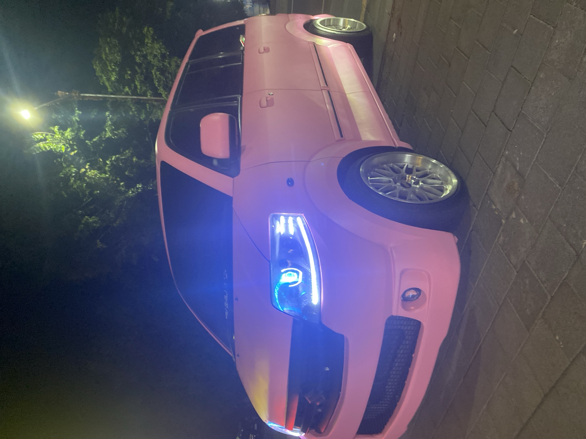 A pink car is parked on a cobblestone street in Unknown, Indonesia at night. The car's headlights are illuminated, and streetlights shine through nearby trees.