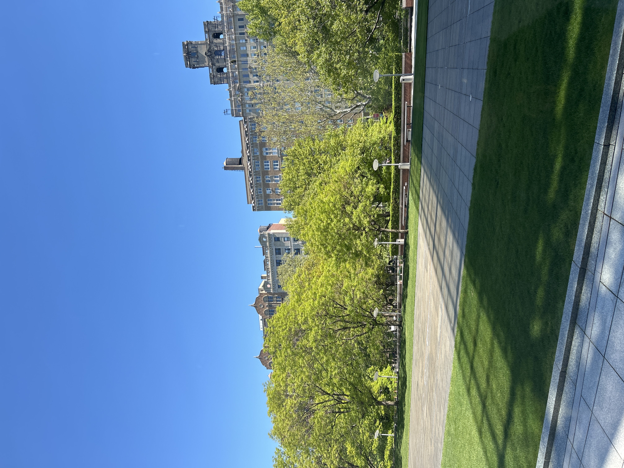 This outdoor shot captures a clear, sunny day in New York City, United States of America. The sky is a vivid blue without any clouds. The foreground features a meticulously maintained park with lush green grass and a stone-tiled walkway.
Tall, mature trees with full, vibrant green leaves line the park, partially obscuring the buildings in the background. These buildings vary in height and architectural style, ranging from older, ornate structures to more modern high-rises. Some buildings are constructed with red brick, and the facade of some buildings is visible.
The scene appears calm and undisturbed, with no immediate presence of people or evident activity. The lighting is bright and natural.