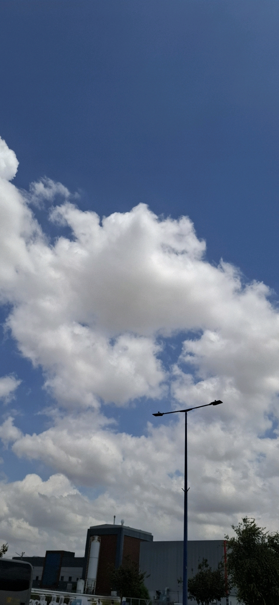 The image shows a bright blue sky filled with scattered white cumulus clouds. In the foreground, a silhouetted lamppost with two lights is prominently visible. Below the lamppost and in the lower portion of the image, there is a view of industrial or commercial buildings. One building features a dark brick facade with lighter colored columns, and a tall white cylindrical silo attached to its side. Another section of the building appears to be clad in gray metal panels. To the left of these buildings, the dark side of a vehicle, possibly a bus, is partially visible. The scene appears to be set in an outdoor, possibly industrial area, under a clear sky with a few clouds. The context suggests this location is in Kiryat Gat, Israel, given the provided location information.