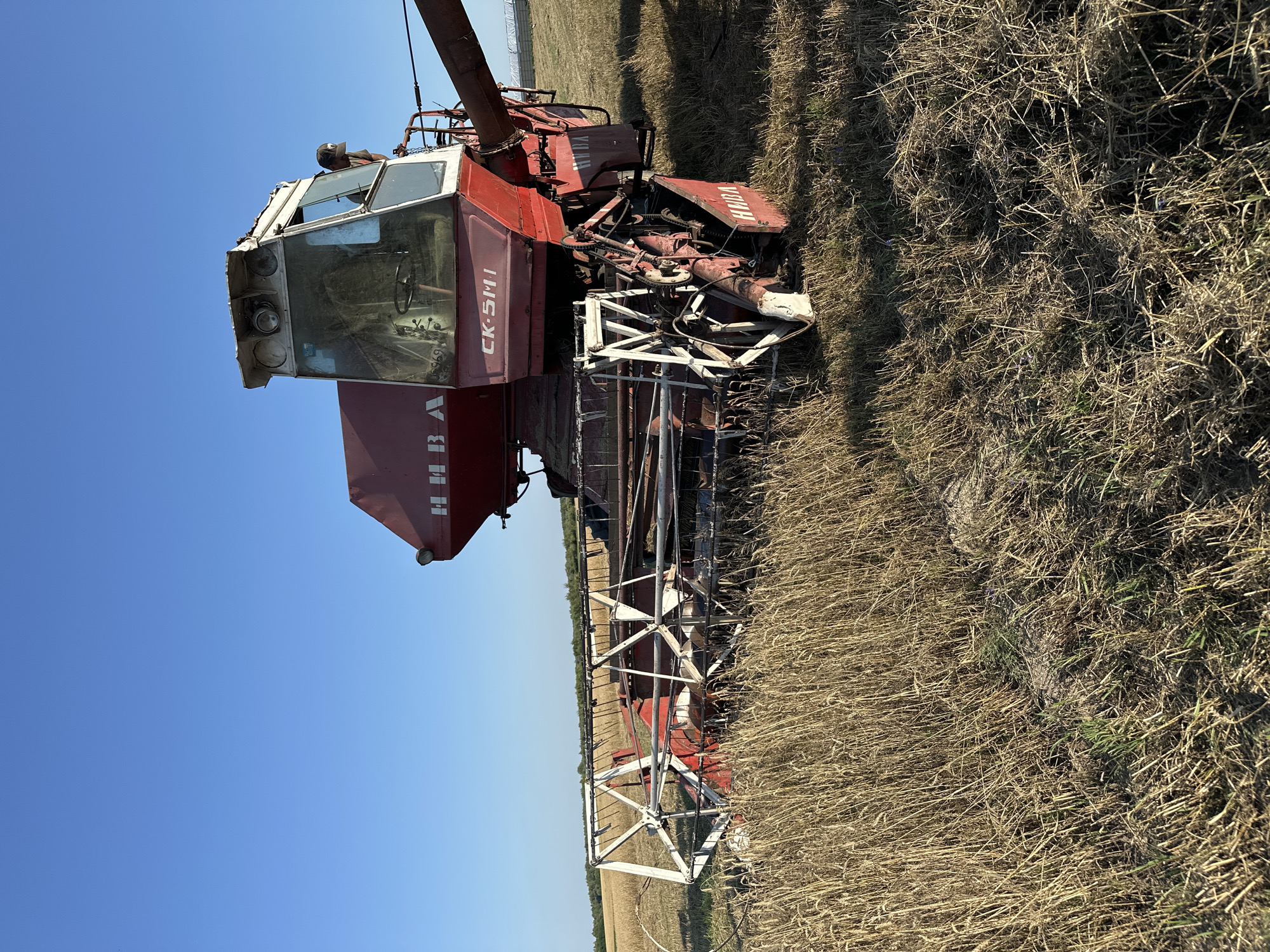 An old, red and white combine harvester is parked in a dry field of wheat, likely in the city of Dubyshche, Ukraine. The combine is positioned at an angle, with its front header resting low in the stubble. The machine appears to be a Soviet-era model, possibly a NIVA SK-5. The cab is visible, with a person wearing a black baseball cap standing in the doorway. The sky above is clear and blue, suggesting a sunny day. The surrounding field is harvested, with remnants of wheat stalks and dry grass. A faint line of trees can be seen in the distance on the horizon.