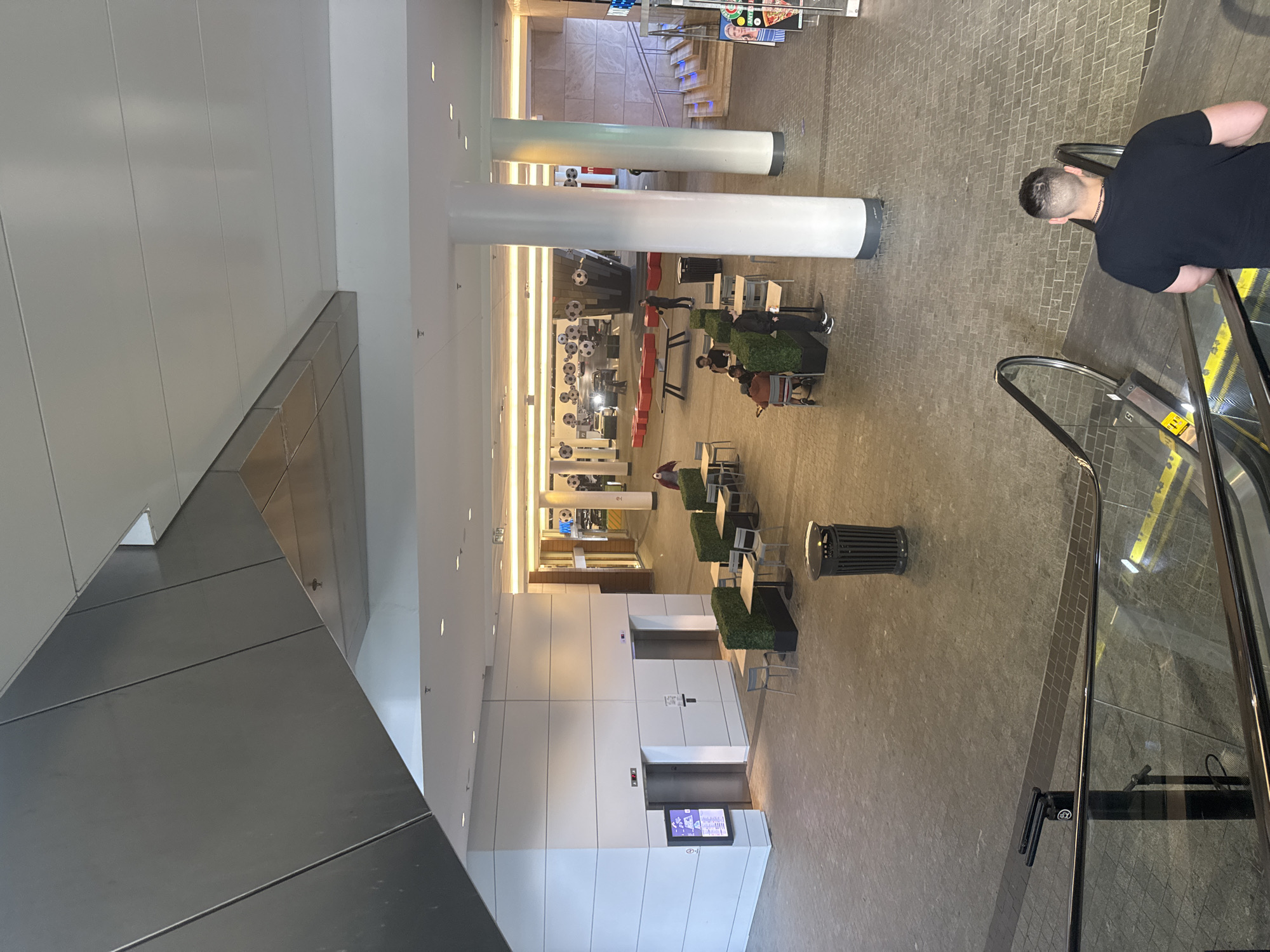This image captures an interior view of the Amazing Food Building at Simon Fraser University in Burnaby, Canada. In the foreground, a man with short hair and a black t-shirt is ascending an escalator. The escalator has yellow safety markings on its steps and a glass railing. Further into the frame, the ground level of the building features a tiled floor and several seating areas with tables and chairs, some surrounded by green planters. Two elevators are visible on a white wall, one with a digital display showing "2FL." A large, grey trash can stands near the seating area. In the background, the space opens up to a wider area with more seating, red tables, and decorative soccer balls hanging from the ceiling. Cars are visible in the background, suggesting this area might lead to a parking or drop-off zone. The architecture includes large white columns and a complex, angled ceiling structure made of metal and white panels. A staircase is visible on the right side, leading to an upper level.