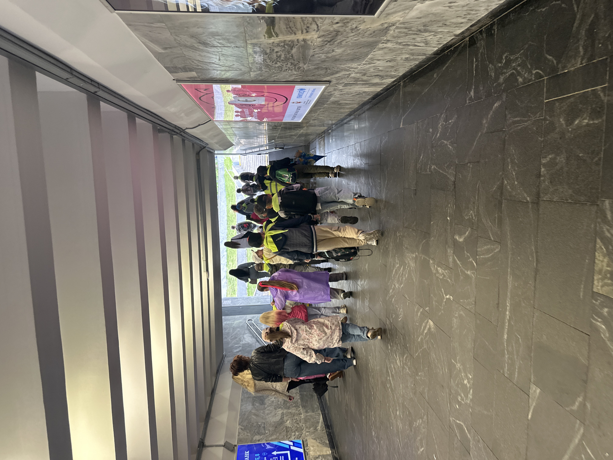 In a pedestrian underpass in Minsk, Belarus, a group of children and adults are walking towards an exit. The underpass is illuminated by fluorescent lights in a ribbed ceiling. The walls are lined with grey marble-patterned panels, and there are posters on the right wall. The floor is tiled with large dark grey stones. The people are walking away from the camera, mostly in the same direction. Some of the children are wearing bright yellow safety vests, and one child is wearing a purple raincoat. A woman in a black leather jacket and jeans is walking on the left, followed by a child in a floral jacket. Another woman with blonde hair is walking behind her. The group appears to be moving through the underpass together.