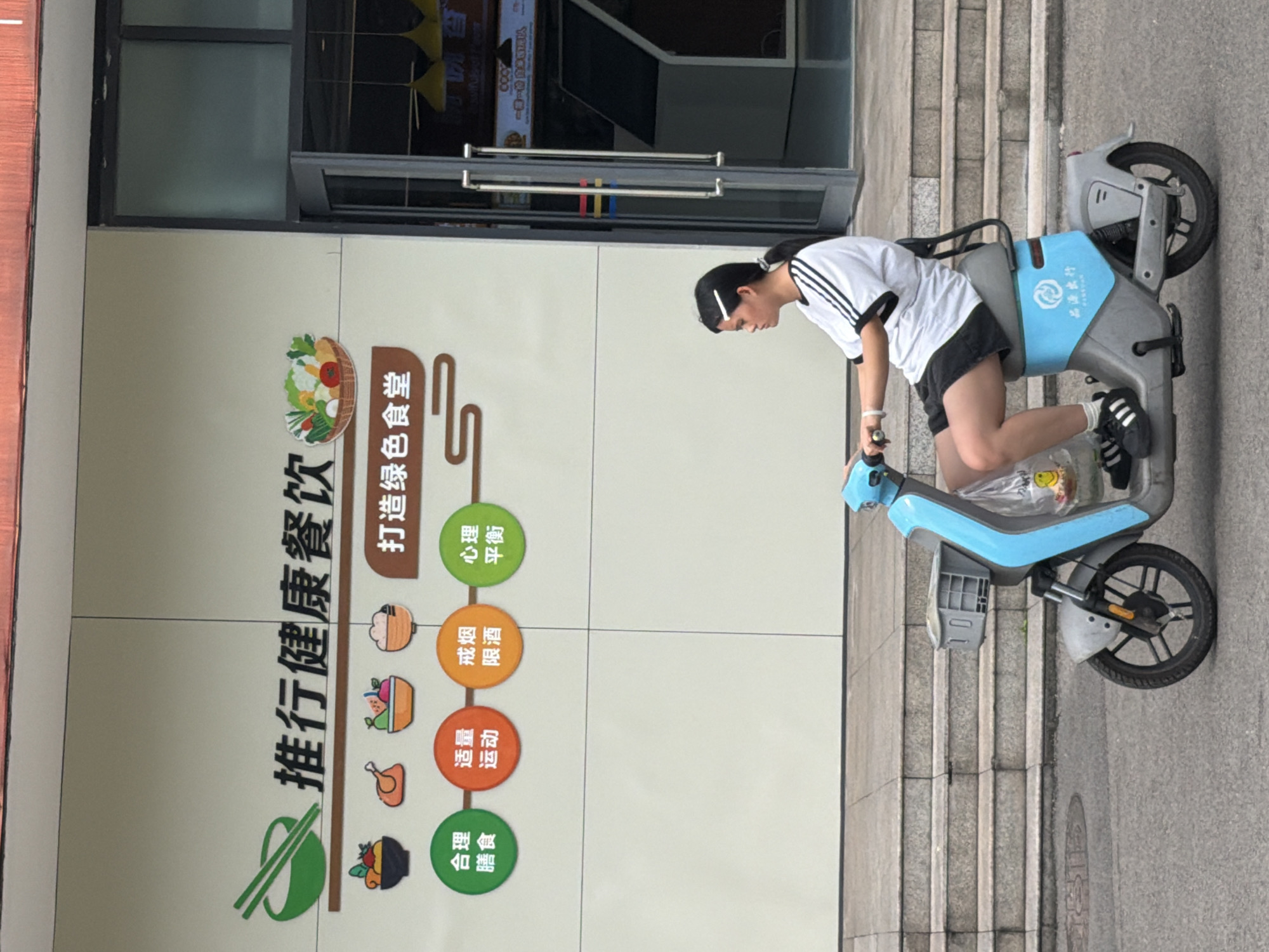 An outdoor, eye-level shot captures a young person interacting with a blue and grey electric scooter on a paved area next to a building. The scene appears to be in Jiangjin, China, given the visible Chinese text.
The person, possibly a female with dark hair tied back, wears a white t-shirt with black stripes on the sleeves, black shorts, and black sneakers with white laces and stripes. She is leaning forward, seemingly adjusting or preparing to ride the scooter, with her right foot on the ground and her left leg bent on the scooter's platform. A plastic bag with some contents is visible between her legs on the scooter.
The electric scooter is light blue and grey. The blue section on the seat area clearly displays the Chinese characters "美团单车" (Meituan Danche), indicating it is a bike-sharing service operated by Meituan.
Behind the person and scooter is a large, light-colored wall with a prominent graphic sign. The sign features Chinese text and illustrative icons related to health and wellness. The main headings read "推广健康餐饮" (Promote Healthy Dining/Catering) and "打造绿色食堂" (Build Green Canteen/Cafeteria). Below these, various icons are paired with health tips:
* A bowl of vegetables/meat next to "合理膳食" (Balanced Diet)
* A chicken leg next to "适量运动" (Moderate Exercise)
* A bowl of noodles/rice next to "戒烟限酒" (Quit Smoking, Limit Alcohol)
* A brain/mind icon next to "心理平衡" (Psychological Balance)
To the right of the wall, a set of concrete steps leads upwards, and the ground surface is concrete. The building behind the wall has large glass doors and windows, reflecting some interior elements. The lighting suggests it is daytime.