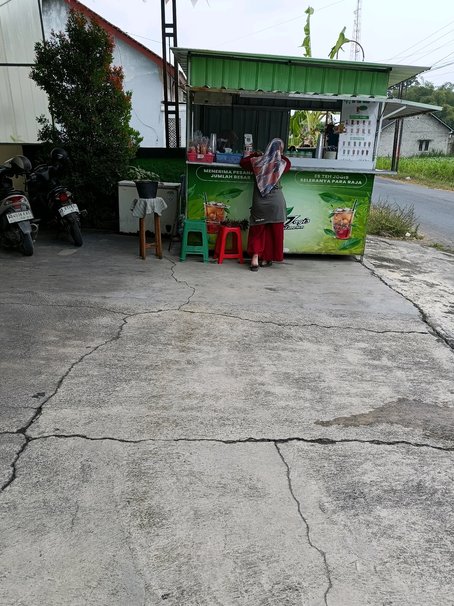 The image shows a street-side food stall in Manisrenggo, Indonesia. A woman wearing a hijab and an apron stands behind the counter, facing away from the camera. She appears to be serving customers or preparing an order. The stall is decorated with green and white banners and features images of iced drinks. Two motorcycles are parked to the left of the stall, and a concrete path with visible cracks stretches in front of it. A residential building and agricultural fields are visible in the background. The sky is overcast, suggesting a cloudy day.