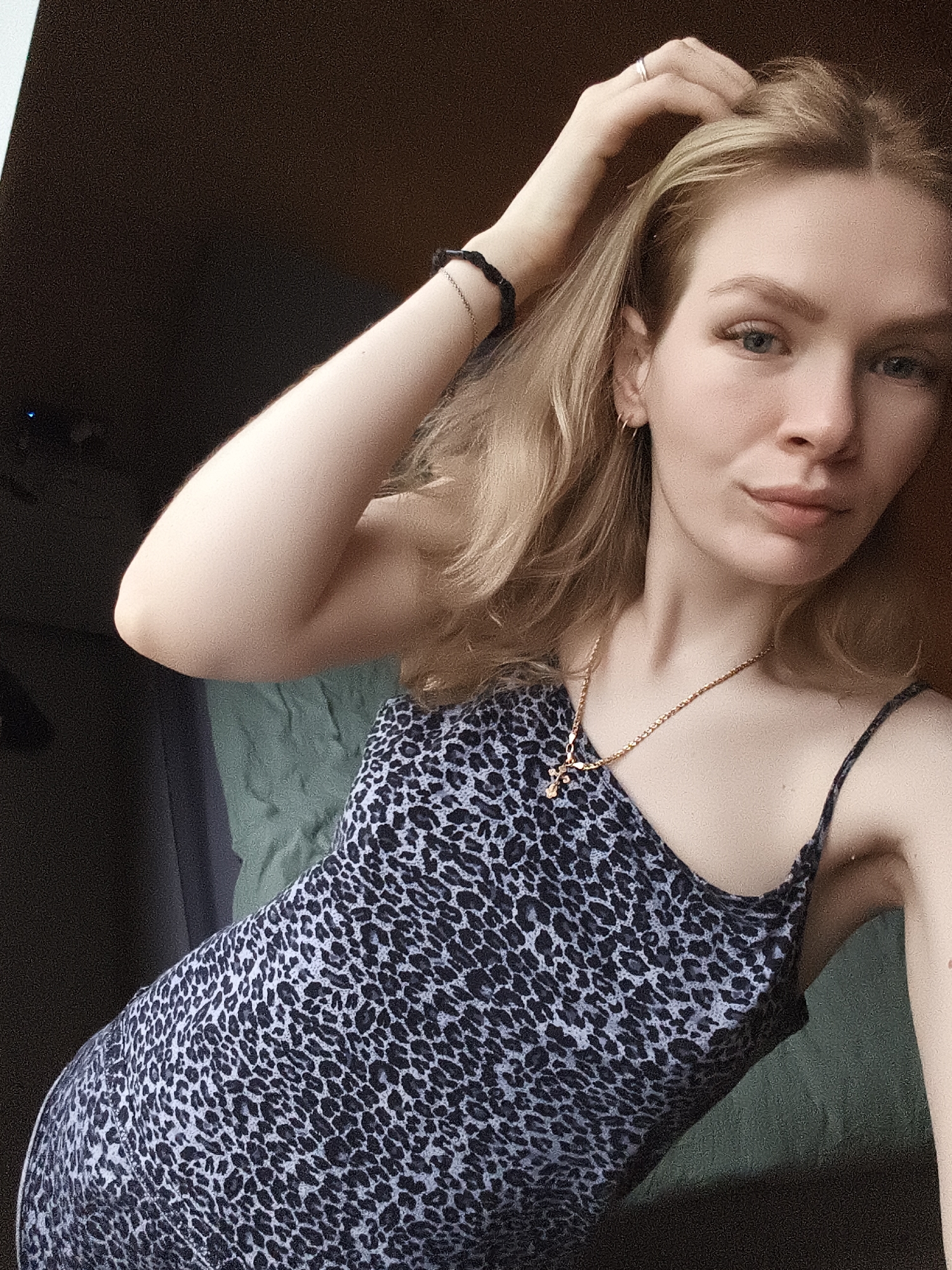 A young fair-skinned woman is in a dimly lit indoor space, possibly in Русский Вожой, Russia. She's wearing a sleeveless dress with a blue and black animal print and a gold chain necklace with a cross pendant. She has blonde hair and blue eyes. Her left hand is raised, touching her hair, and she is wearing a black bracelet on her left wrist and a ring on a finger of her left hand. The background is indistinct, with what appears to be a green blanket or sheet partially visible behind her. The scene suggests an informal, self-portrait style image.