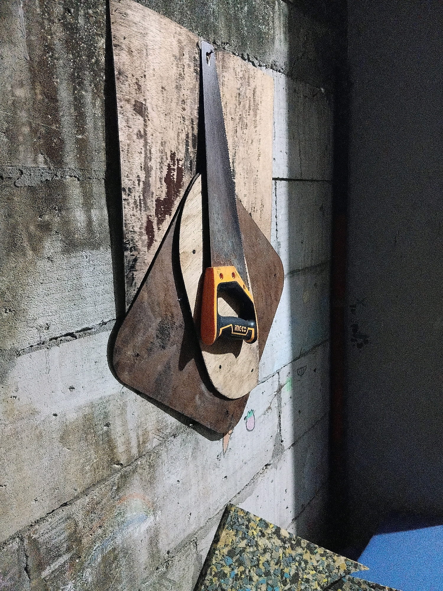 In Cikal, Indonesia, this image depicts a close-up view of a wooden-handled saw hanging on a rough, textured concrete wall. Two pieces of plywood are attached to the wall, serving as a backing for the saw. The larger, darker plywood piece is diamond-shaped, while a lighter, rectangular piece is positioned above it. The saw's blade is visible, and its orange and black handle is prominent. The wall exhibits signs of wear, with patches of discoloration and moss. A shadowed area dominates the right side of the frame, suggesting an indoor or covered space. In the foreground, a piece of foam material with a speckled pattern is visible, along with a sliver of blue surface, possibly a table or floor.