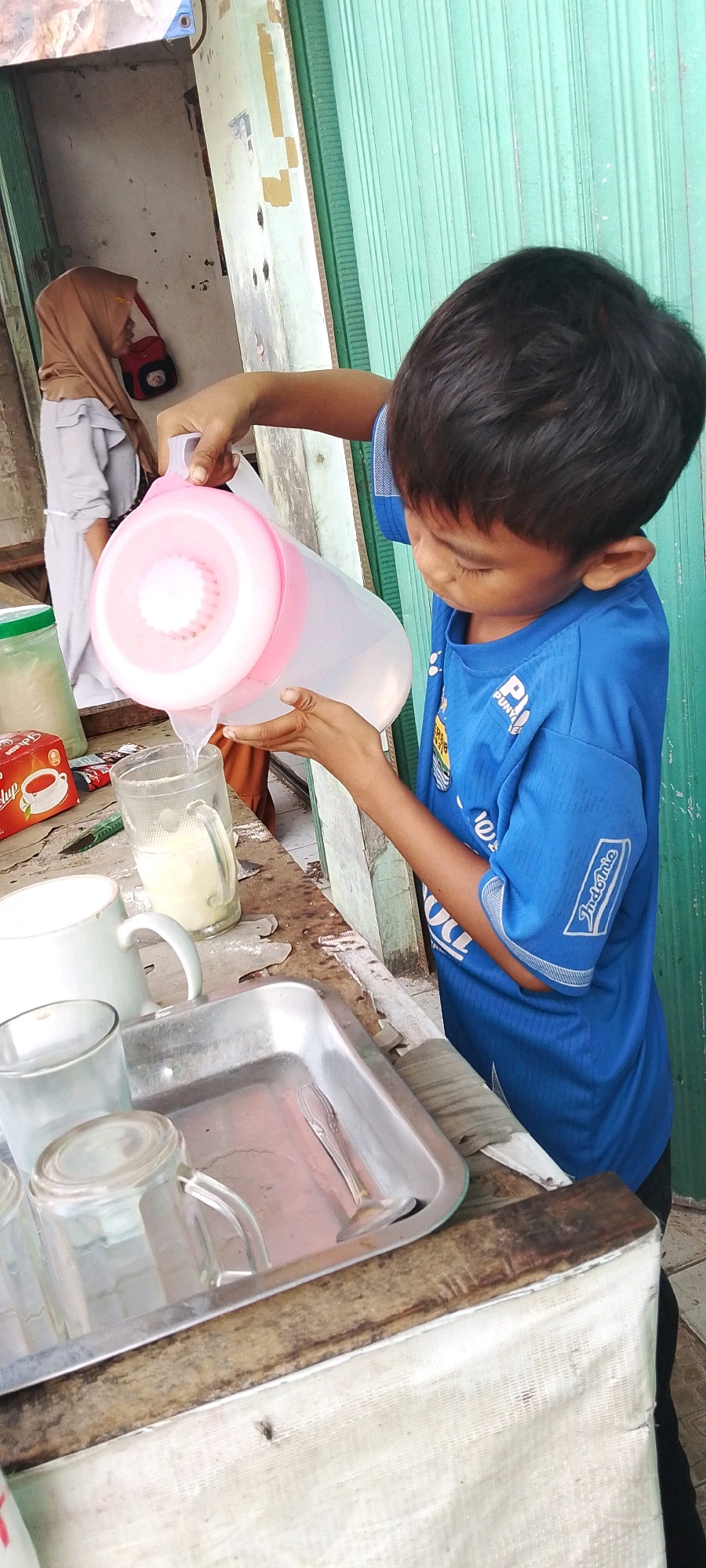 In Sriamur, Indonesia, a young boy in a blue t-shirt is pouring water from a large pink-lidded pitcher into a glass. The pitcher has a filter in its opening. Next to the glass is a box of tea. The boy is standing at a wooden counter that holds several other glasses and a metal tray with a spoon. In the background, a woman wearing a hijab is visible, partially obscured by the counter. The setting appears to be an outdoor vendor stall, with a green corrugated metal door in the background.
