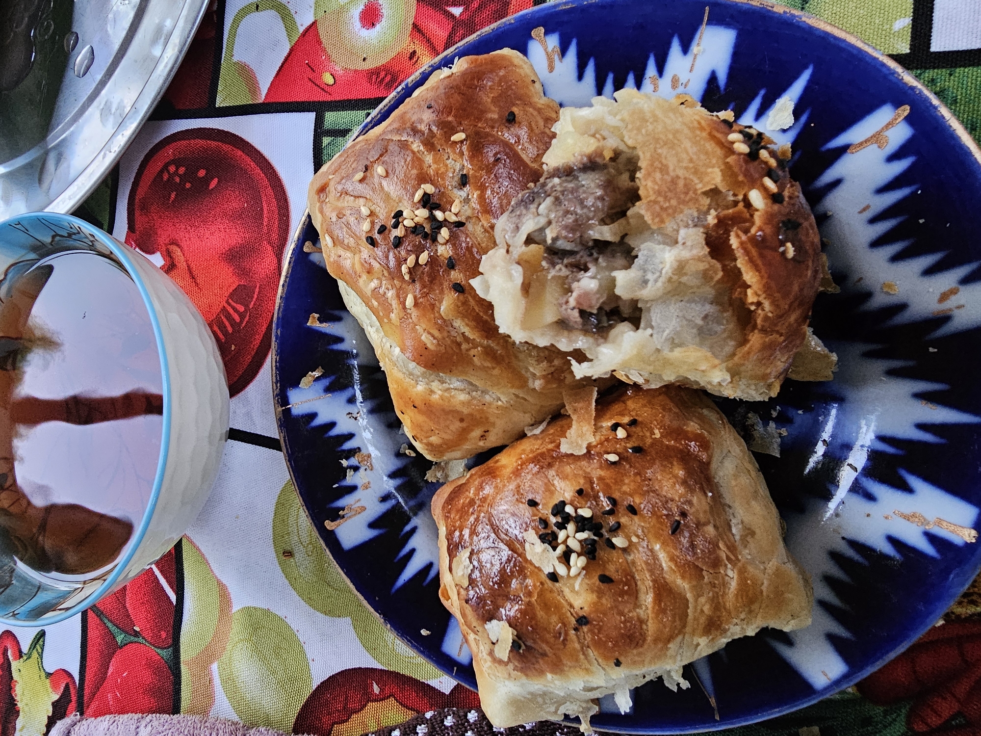 A close-up, overhead shot shows a blue and white patterned plate holding three savory pastries, likely samsa, a popular dish in Central Asia. Two pastries are intact, golden brown with a flaky crust, sprinkled with white and black sesame seeds. The third pastry is partially broken, revealing a filling of minced meat and possibly onions or potatoes. The plate rests on a colorful tablecloth featuring stylized red apples and green and yellow fruit shapes. To the left of the plate, a white ceramic bowl filled with a dark amber liquid, possibly tea, is visible. A pink towel peeks into the frame on the far left. In the background, a metallic tray with water droplets is slightly visible. The overall scene suggests a casual meal setting, possibly in Özgön, Kyrgyzstan, a city known for its rich culinary traditions and historical significance.