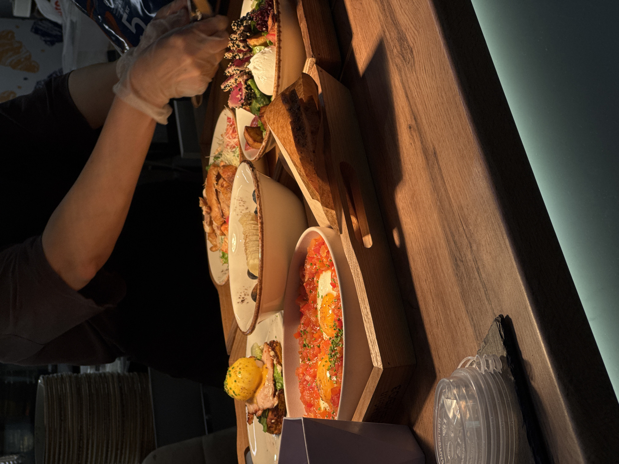A restaurant setting in Moscow, Russia is depicted in the image. The primary focus is on a wooden display stand that features a variety of plated dishes. A person wearing gloves is preparing or arranging the food. The dishes include salad, fried eggs, and what appears to be seared tuna. A stack of clear plastic lids sits on a counter near the wooden stand. The environment features a mix of natural and artificial light.