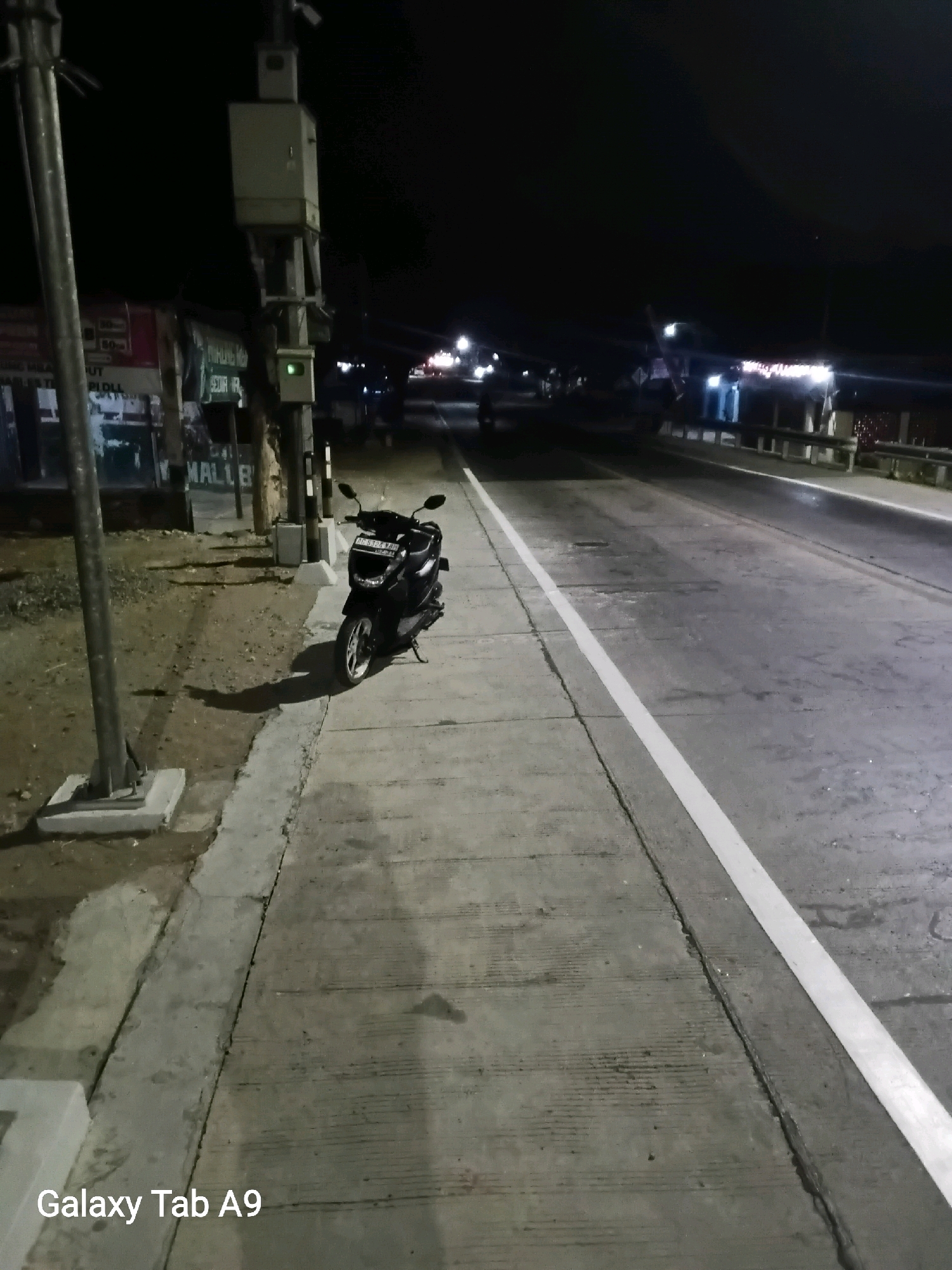 A black motorcycle is parked on a concrete sidewalk next to a road at night. The motorcycle faces left and is on its kickstand. A traffic signal pole with several boxes is behind the motorcycle, to its left. The road has a white line marking the right lane. In the background, lights from buildings and other vehicles are visible. The scene appears to be in a developed area, possibly near businesses. The text "Galaxy Tab A9" is visible in the bottom left corner of the image.