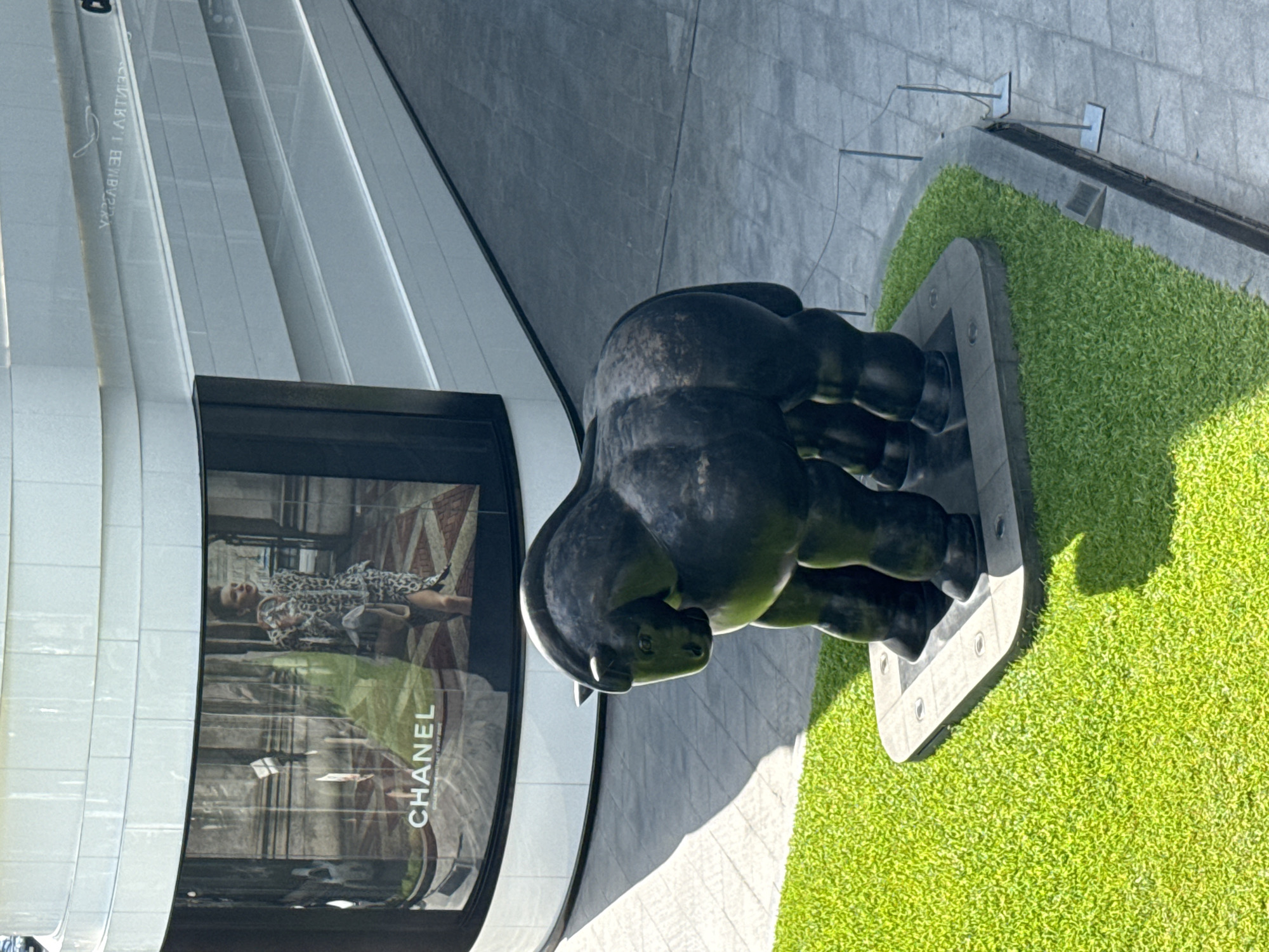 This is a high-angle photograph of a bronze horse sculpture located on artificial turf in front of a modern building with a curved glass facade displaying a "CHANEL" advertisement. The sculpture is dark bronze, with a heavy, stylized form, and stands on a square base. The building features white horizontal paneling and a curved glass window that reflects the surroundings and displays a fashion advertisement. The scene is located in Bangkok, Thailand, as indicated by the building's architecture and the overall style. The lighting suggests it is daytime, casting shadows from the sculpture onto the turf and pavement.