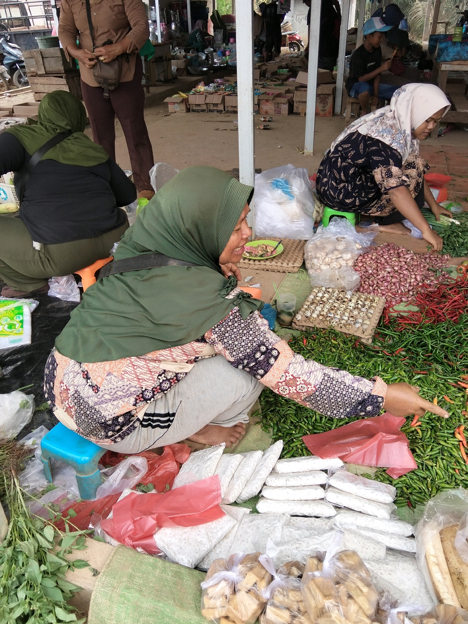 A group of women are selling produce at an outdoor market in Tirto Sari, Indonesia. In the foreground, a woman wearing a green hijab and a patterned shirt is pointing to a pile of green chilies. She is sitting on a blue stool. Next to her is another woman wearing a black top and green pants, also wearing a green hijab. In the background, a woman in a white hijab and patterned shirt is crouched down by a pile of green onions. Other women are visible in the background, as well as stalls and produce. The market appears to be bustling with activity.