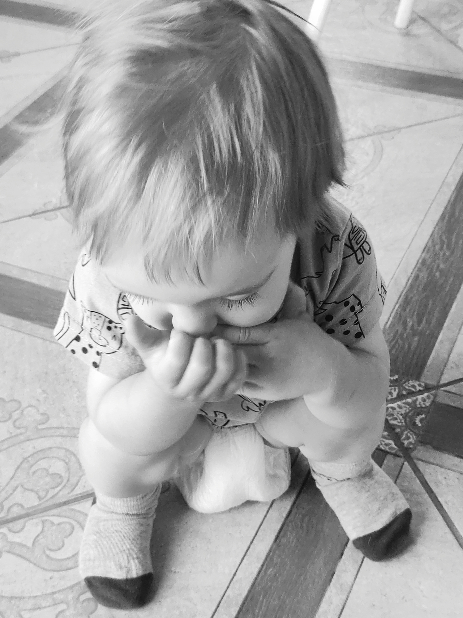 A baby with blond hair sits on a tiled floor. The baby wears a shirt with cartoon figures and socks with dark toes. The baby's hands cover the mouth. The background is a tiled floor and a blurred chair. The location is not specified in the image.