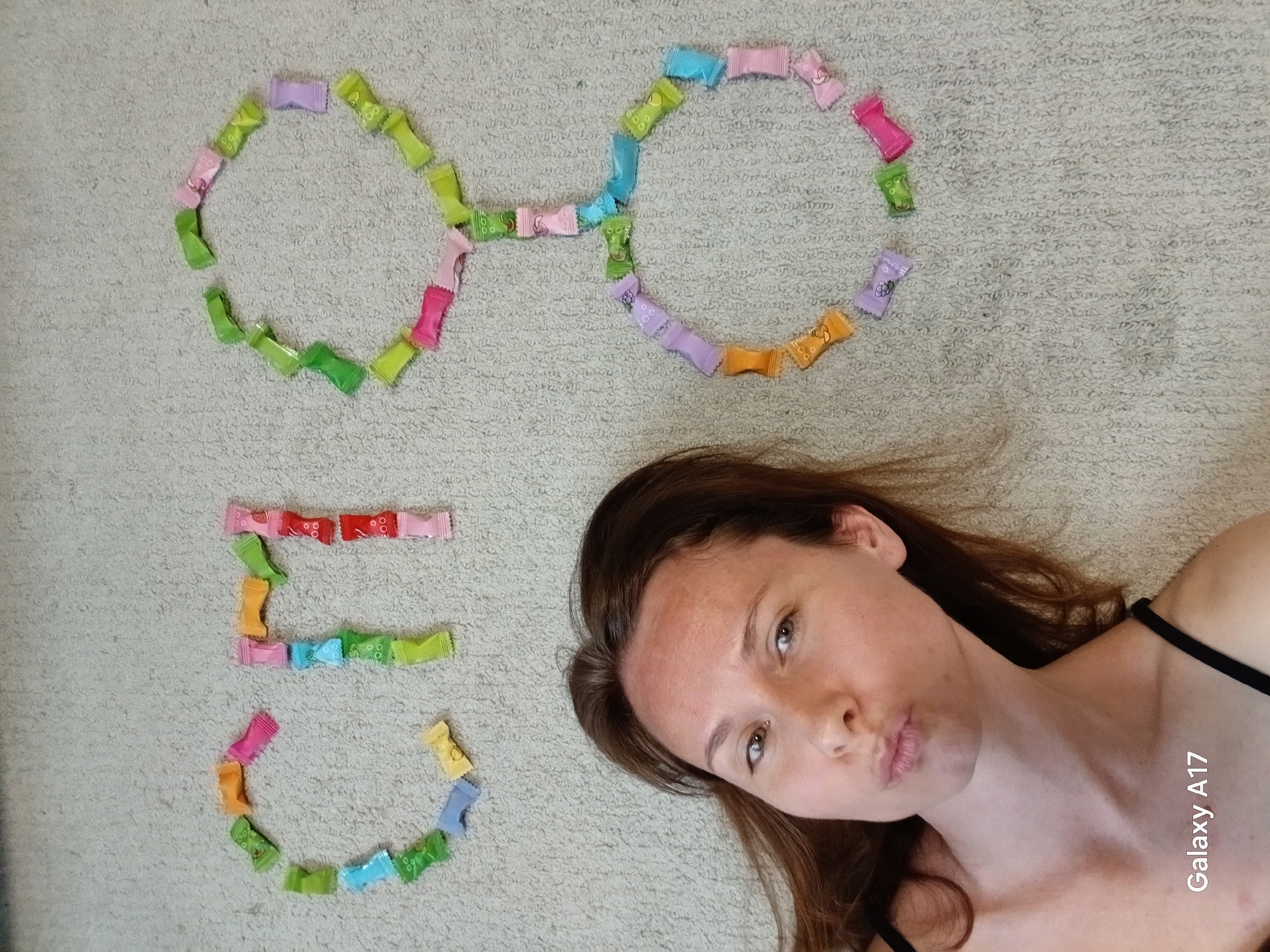 A young woman with brown hair and light skin is lying on a beige carpet, looking towards the camera with pursed lips. To the left of her head, the number "50" is formed with small, colorful, rectangular candies. Above the number, a pair of eyeglasses is created from the same candies. The photograph seems to be taken indoors with natural lighting. The bottom right corner of the image displays the text "Galaxy A17." There is no information in the image suggesting the location of Yekaterinburg, Russia.