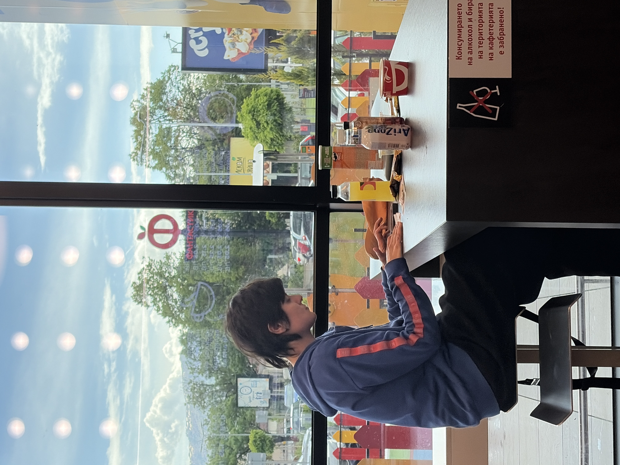 A person is sitting at a table inside a cafe, looking out the window. The cafe's interior is visible, including a table with drinks and snacks, and a sign prohibiting alcohol consumption in Bulgarian. Outside the window is a view of a street in Sofia, Bulgaria. Visible in the scene are an Arizona Iced Tea can and Pringles container.