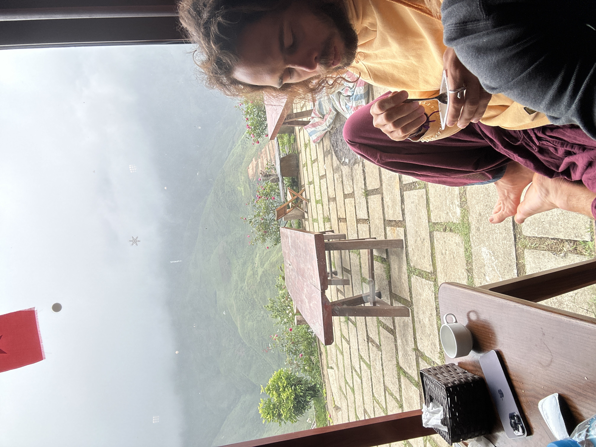 A young man with curly brown hair and a beard, wearing a yellow hoodie and maroon pants, is sitting on a tiled patio. He is holding a small white container and a spoon, appearing to be eating. His eyes are closed and he is looking down. In the foreground, a wooden table holds a black napkin holder, a white ceramic mug, and a smartphone. Beyond the patio, a lush green mountainous landscape is visible through a window, shrouded in mist. The scene suggests a peaceful, contemplative moment in a natural setting. The location is identified as Ta Xua, Vietnam.