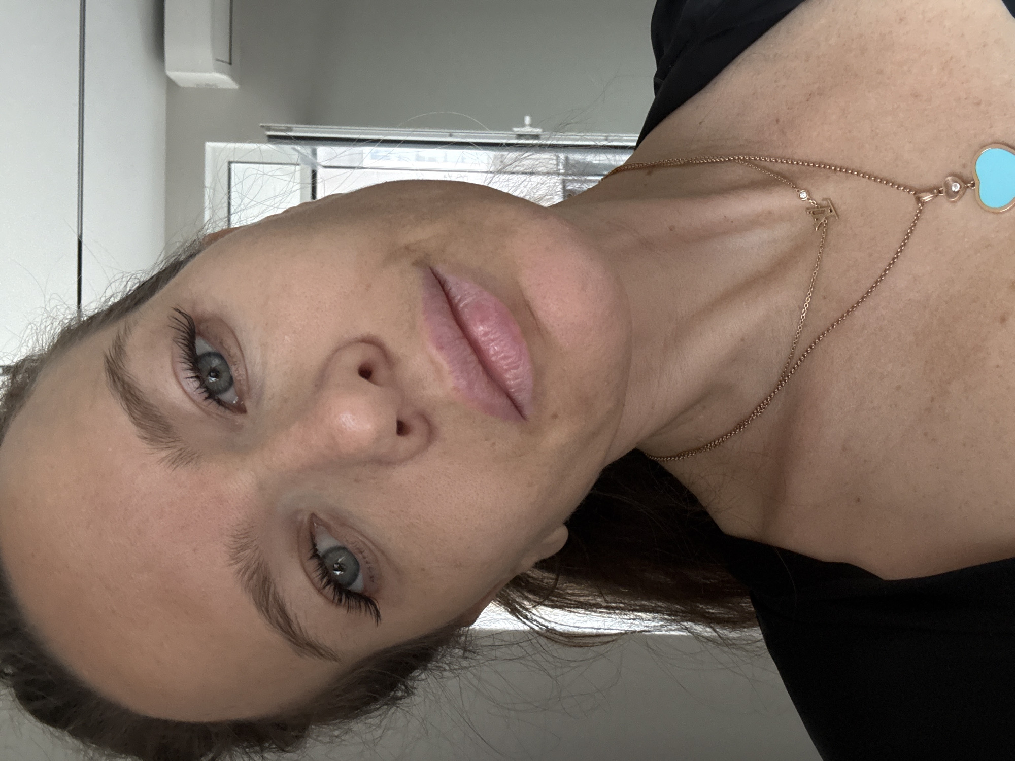 A close-up, angled self-portrait of a woman with light skin, gray-blue eyes, and brown hair is presented. She has a gold necklace featuring a Tiffany blue heart pendant and a diamond "LV" logo. She is wearing a black top. The background includes a partially visible window with white frames and a white wall, possibly indoors. There is no indication of Dolgoprudny, Russia in the image.