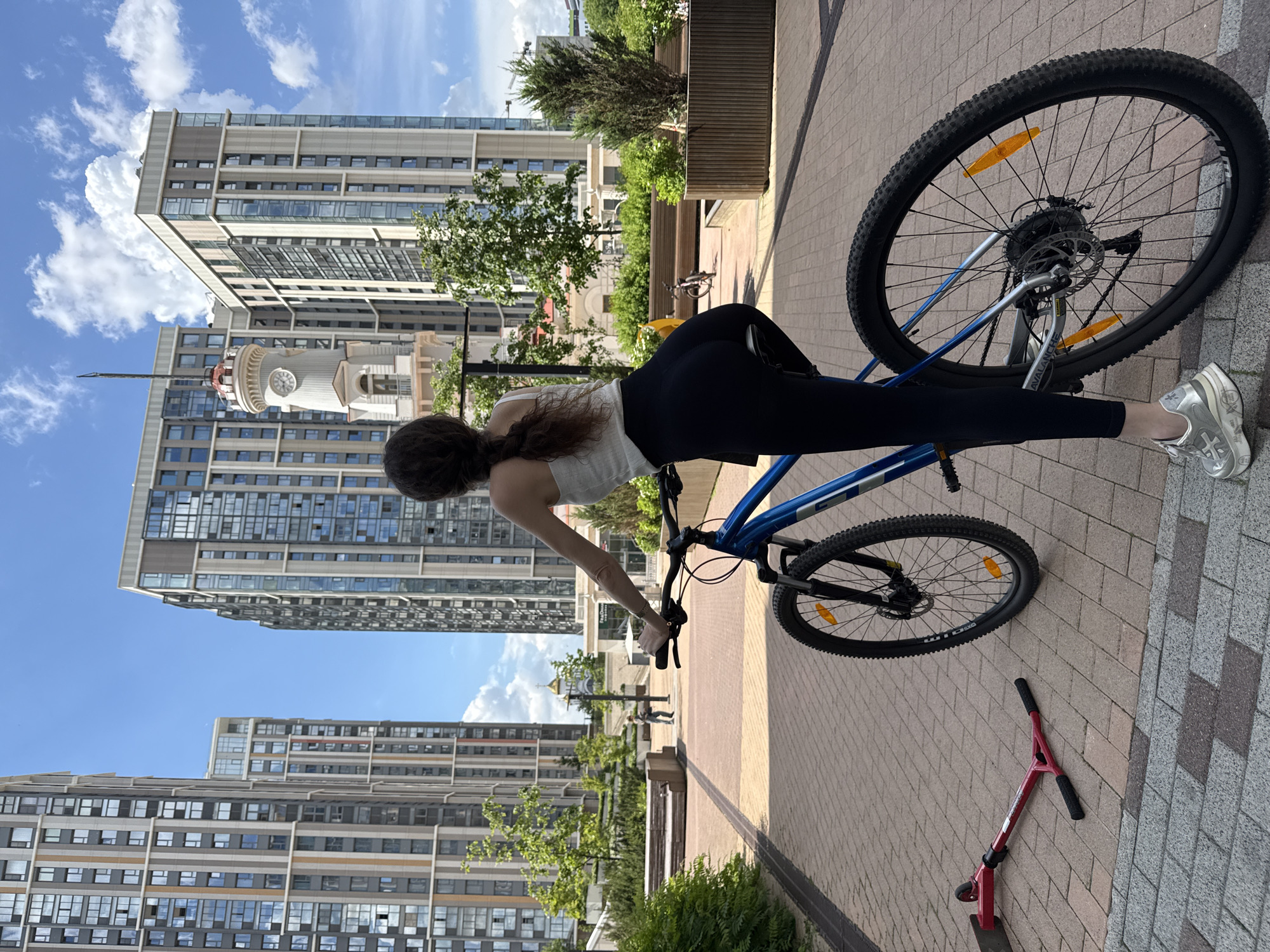 An outdoor, eye-level shot captures a young woman and her bicycle in a modern urban setting under a clear blue sky. The image is oriented vertically, with the woman standing to the left, facing away from the camera. She has long, dark brown hair in a braid and is wearing a white tank top and black leggings. Her body is bent over a blue and silver mountain bike, with her right leg extended towards the rear wheel, suggesting she is adjusting or interacting with the bike.
The bicycle, a GT Avalanche model with "MAXXIS" tires and disc brakes, is positioned prominently in the mid-ground. Next to the bike, a red scooter with "K" and "O" letters partially visible on its frame rests on the paved ground. The ground is a mix of light brown and gray brick patterns.
In the background, several tall, contemporary apartment buildings with numerous windows rise into the sky, reflecting the sunlight. One building features a distinctive clock tower with a dome and a spire, its clock face showing approximately 10:10. Green trees and bushes are interspersed throughout the scene, along with wooden benches, indicating a pedestrian-friendly area. The bright sunlight and scattered white clouds suggest a pleasant daytime, likely morning or early afternoon. The architecture and layout are characteristic of modern residential complexes found in cities like Moscow, Russia.