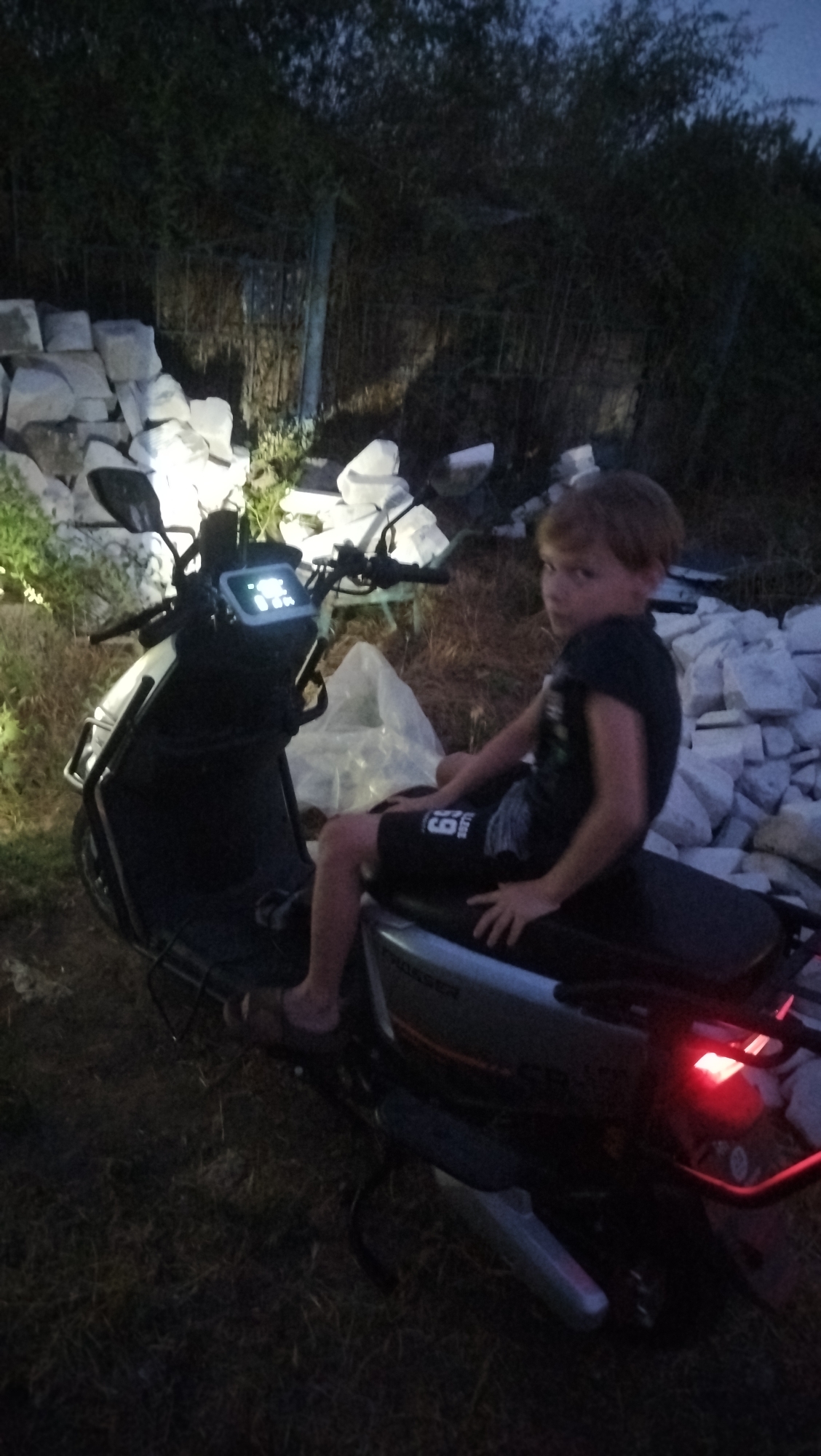 A young boy with blond hair sits on a parked electric scooter in a dimly lit outdoor area, possibly Kostiantynivka, Ukraine. The boy is wearing a black t-shirt with a graphic design and black shorts with the number "69" printed on them. He is looking towards the camera with a neutral expression. The scooter is dark in color with a digital display illuminated on the handlebars. In the background, there are piles of light-colored stones and dense vegetation. The overall lighting suggests it is dusk or night.