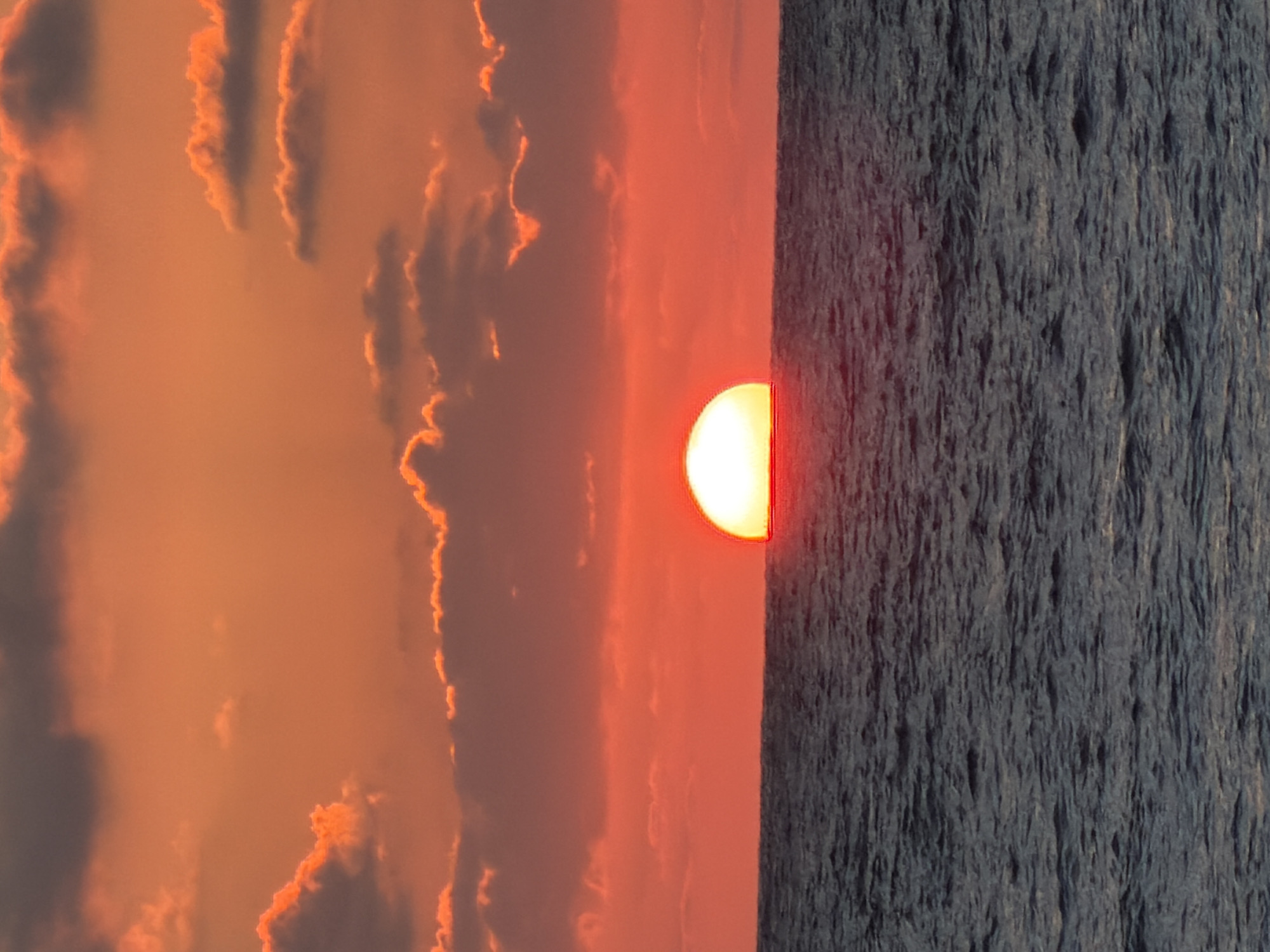 The image depicts a sunset over the Mediterranean Sea, likely taken in or near Tel Aviv, Israel. The sun is bisected by the horizon line, with the lower half reflecting intensely on the water's surface. The sky is a blend of orange and red hues, with clouds edged in a lighter orange. The sea is dark blue/gray, with a textured surface indicating wave activity. There are no people or specific objects visible other than the sun, sea, and clouds. The overall impression is one of a natural sunset scene.