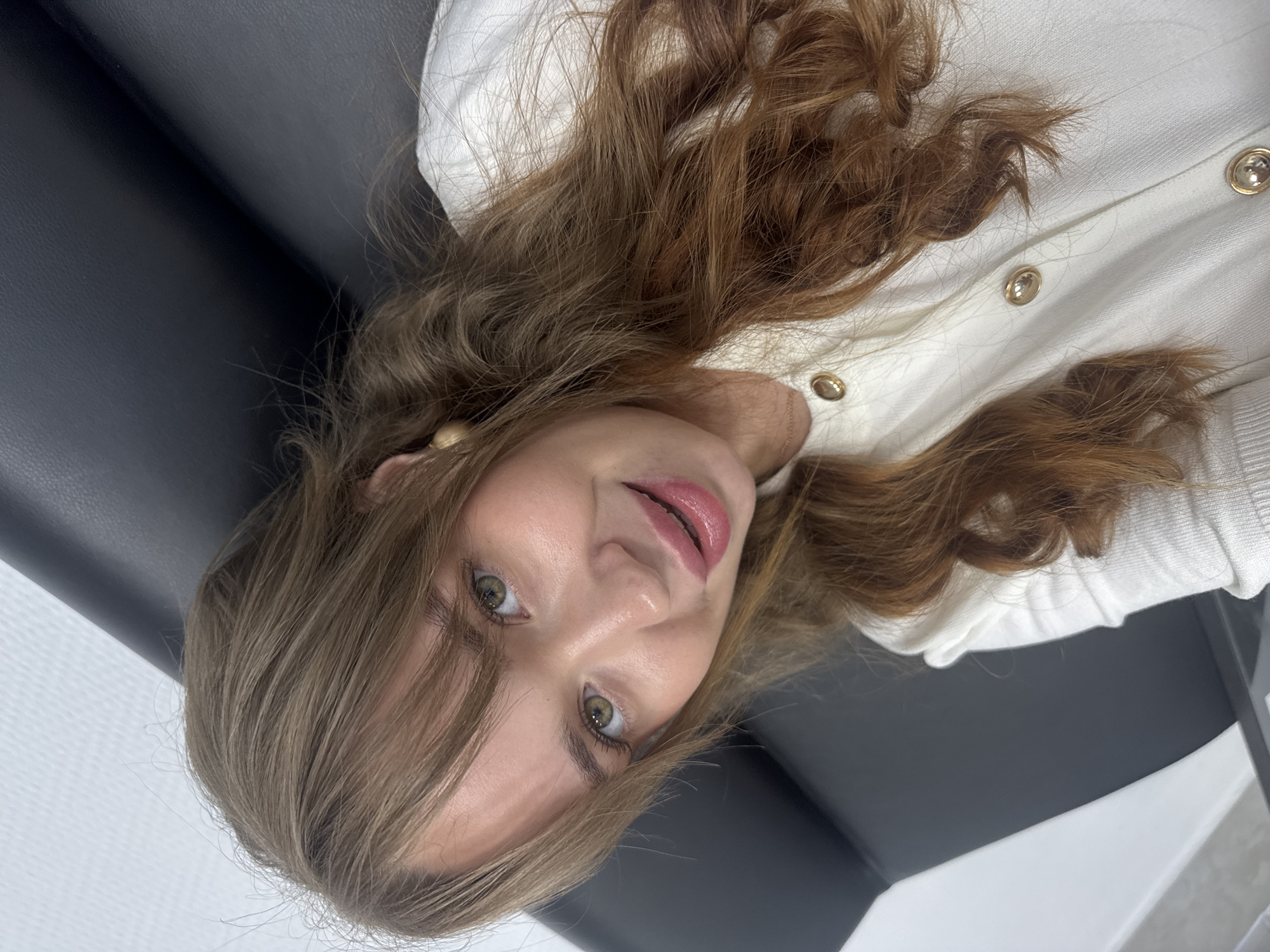 A medium close-up photo depicts a young woman with long, wavy, light brown and auburn hair, lying down and looking directly into the camera. The image is oriented with her head on the left and her body extending to the right. She has green eyes, fair skin, and her lips are slightly parted, revealing a pink gloss.
She is wearing a light-colored (likely white or off-white) knitted cardigan or sweater adorned with gold-colored buttons down the front. A gold earring is visible on her left ear, and a thin, gold-colored chain or bracelet is partially visible on her right wrist, near her neck.
The woman is positioned on a dark, smooth surface, which appears to be upholstery, possibly a couch or chair. A lighter, white surface is visible behind her upper body, suggesting an indoor setting. The background is simple and slightly out of focus. Her expression is calm and engaging, with a hint of a smile. There are no specific activities taking place other than posing for the photograph. No text is visible in the image. The location context is Chaikovsky, Russia.