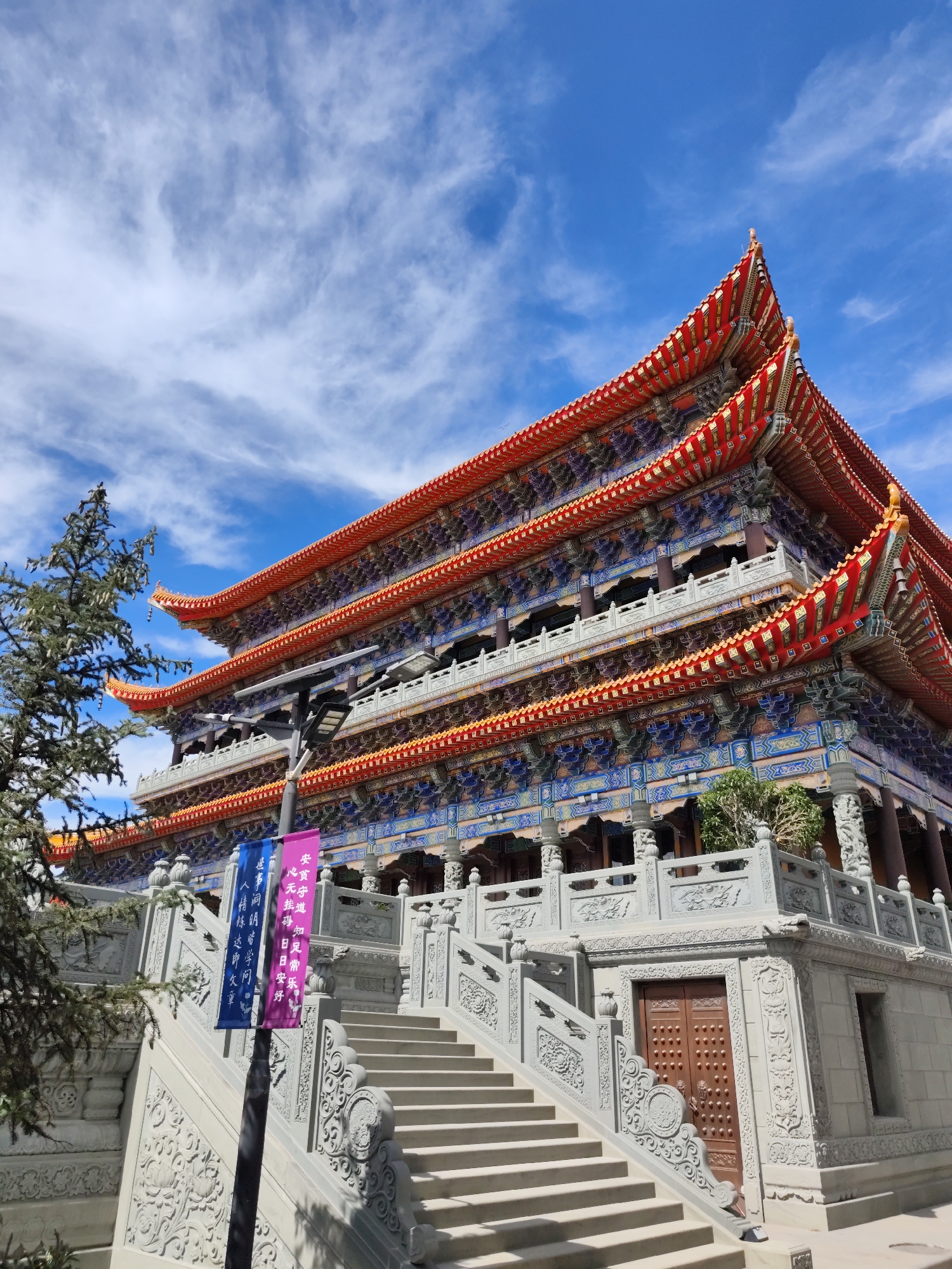 A multi-tiered, ornate Buddhist temple with red and gold roofs and intricately painted blue and gold details on its facade is seen under a bright blue sky with wispy white clouds. The temple is approached by a wide, stone staircase with carved railings and decorative elements. A tall lamppost with two hanging banners, one blue and one pink, stands to the left of the stairs. A pine tree with dark green foliage is visible on the far left of the image. The temple appears to be located in the city of Daqing, China, specifically at the Daqing Tibetan Buddhist Temple. There are no people visible in the image. The scene is static and depicts an architectural landmark.