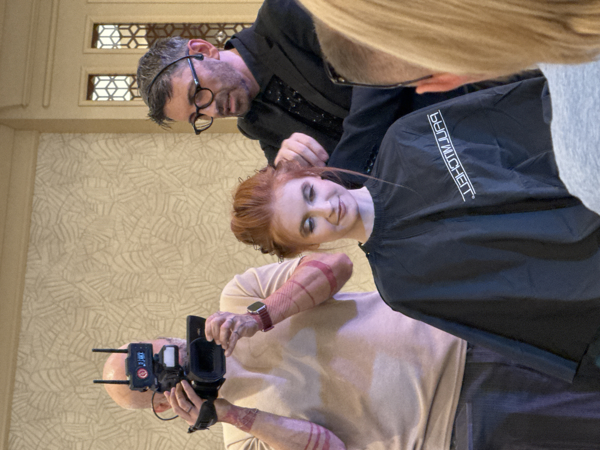 The image shows a man with glasses wearing a black blazer, working on a woman's red hair, who is wearing a black salon cape with white writing on it. Another person, possibly a photographer, is capturing the moment using a camera. The environment seems to be indoors with patterned wallpaper. The location is Unknown, Unknown.