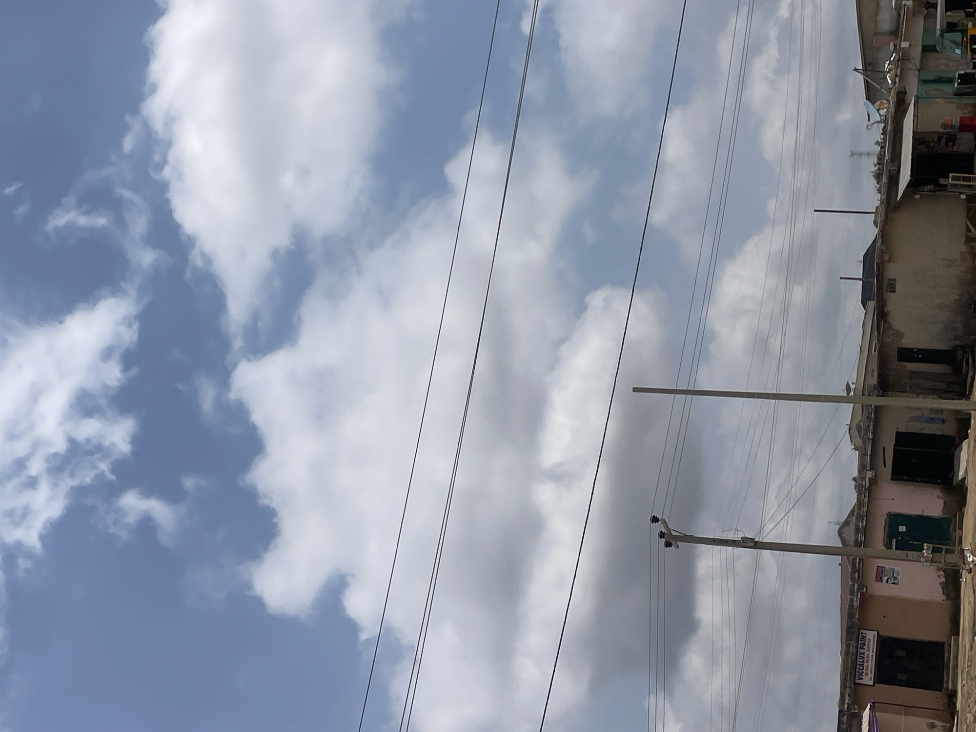 This media file depicts a street scene in Unguwar Saya, Nigeria, characterized by a partly cloudy sky. The image is oriented vertically, with utility lines and a power pole prominently featured in the foreground, stretching across the frame. These lines are suspended against a backdrop of blue sky interspersed with fluffy white clouds.
To the right of the utility lines, a row of buildings lines the street. These structures appear to be shops or residences, with some exhibiting signs of wear and tear. One building has a sign that reads "VICCALUX PAINT." The architecture suggests a typical urban or semi-urban environment in Nigeria. The lighting indicates it is daytime, likely late morning or early afternoon, given the brightness and the nature of the shadows. The weather appears fair, with the clouds suggesting potential for shade but not an imminent downpour. There are no people visible in the frame, focusing the viewer's attention on the built environment and infrastructure. The overall impression is one of everyday life in a Nigerian settlement, with a focus on the infrastructure supporting it.