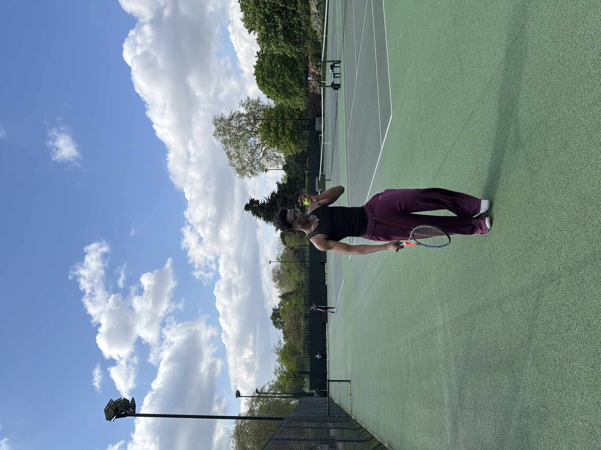 The image, rotated 90 degrees clockwise from its correct orientation, captures an outdoor tennis scene under a bright, partly cloudy sky. In the foreground, a person is depicted mid-serve, holding a yellow tennis ball aloft with one hand and a tennis racket with an orange and blue handle in the other. They are dressed in a black tank top, dark maroon or plum-colored wide-leg trousers, and white sneakers. They have dark, curly hair and appear to be wearing glasses or sunglasses.
The setting is a green tennis court with clear white lines. A tennis net stretches across the court in the mid-ground, beyond which other players are visible, suggesting a game or practice session. The background features abundant green foliage from numerous trees, implying a park or sports club environment. A dark chain-link fence runs along the side of the court, and tall light poles equipped with floodlights stand against the sky.
The scene takes place during the daytime, evidenced by the bright sunlight, clear blue sky, and scattered white clouds. Shadows are visible on the court. There is no discernible text within the image. The location is contextualized as London, United Kingdom.