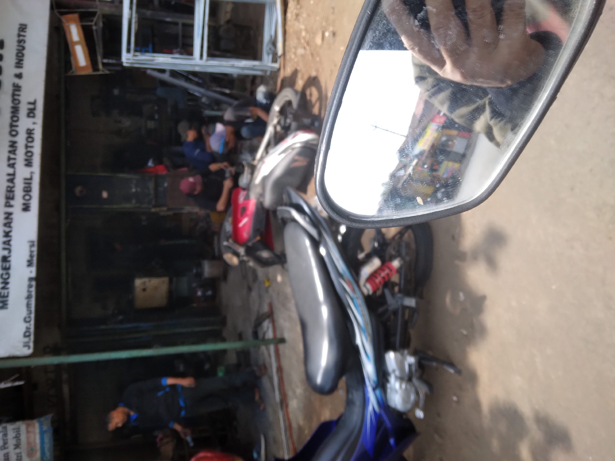 The image depicts a street scene in Sokaraja, Indonesia, likely in front of an automotive repair shop. In the foreground, a motorcycle is partially visible, with its blue and silver body and black seat in focus. A side mirror of another vehicle, possibly a motorcycle, reflects the scene. In the background, the shop appears to be open-air, with various tools and equipment, including what looks like metal frames, scattered around. Several individuals are present, some appearing to be working on vehicles. One person in the middle ground, wearing a dark blue shirt, is bent over a motorcycle. Another person, wearing a dark shirt and a maroon cap, is seated and engaged in some activity. A third person, in a black shirt and blue cap, is also working. The ground is dirt and gravel. A sign above the shop reads "MENGERJAKAN PERALATAN OTOMOTIF & INDUSTRI" and "JL.Dr.Gumbreg - Mersi MOBIL, MOTOR, DLL", indicating the business specializes in automotive and industrial equipment repair, located on Dr. Gumbreg Street in Mersi.