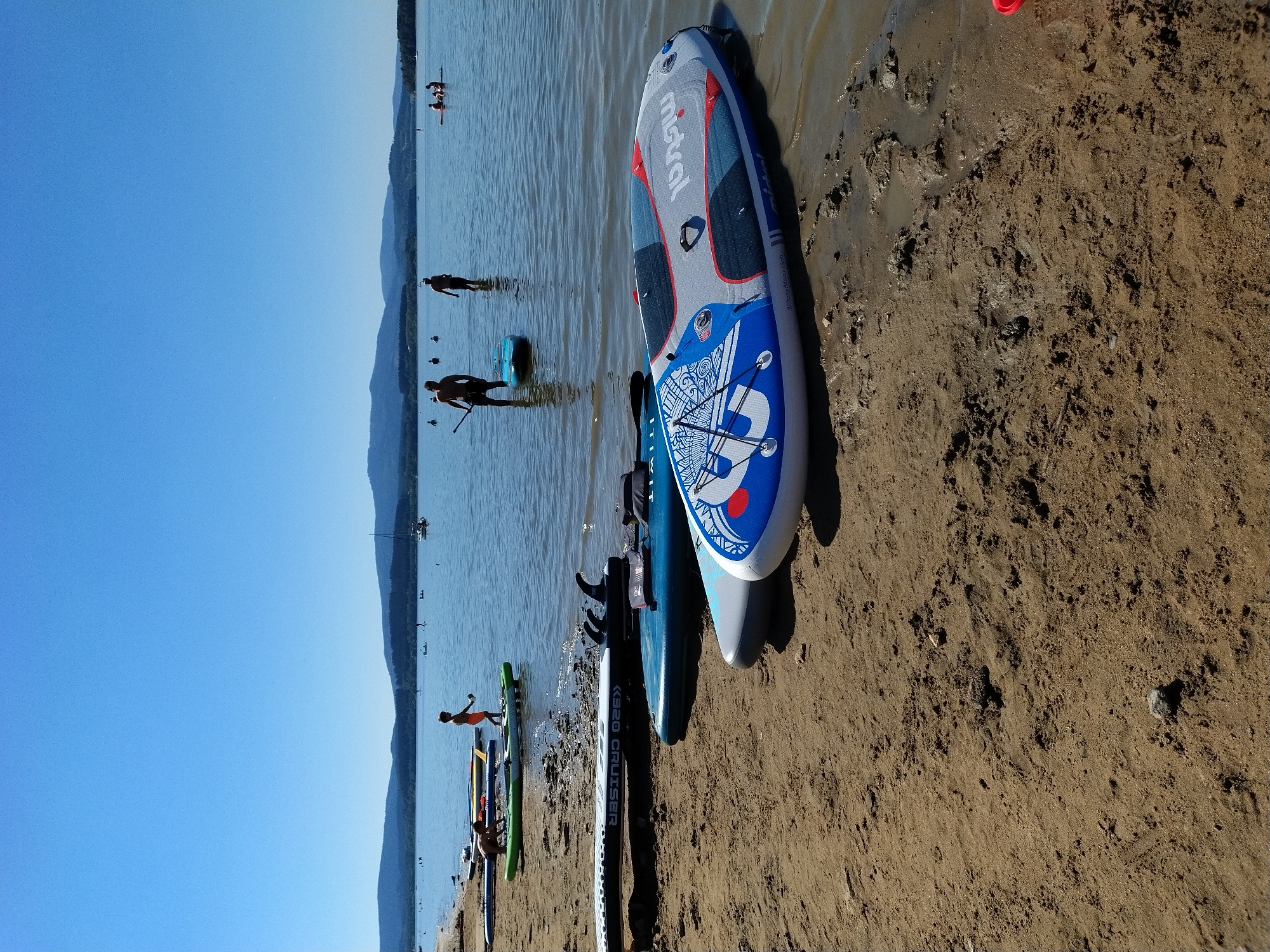 The image depicts a sunny day at a lakefront in Radslav, Czechia. Several people are visible in the water, some standing and others on paddleboards. In the foreground, on the sandy shore, are numerous paddleboards of various colors and designs. The water is calm, and the sky is clear blue with distant hills forming the horizon. The atmosphere appears recreational, with people enjoying water activities on a summer day.
