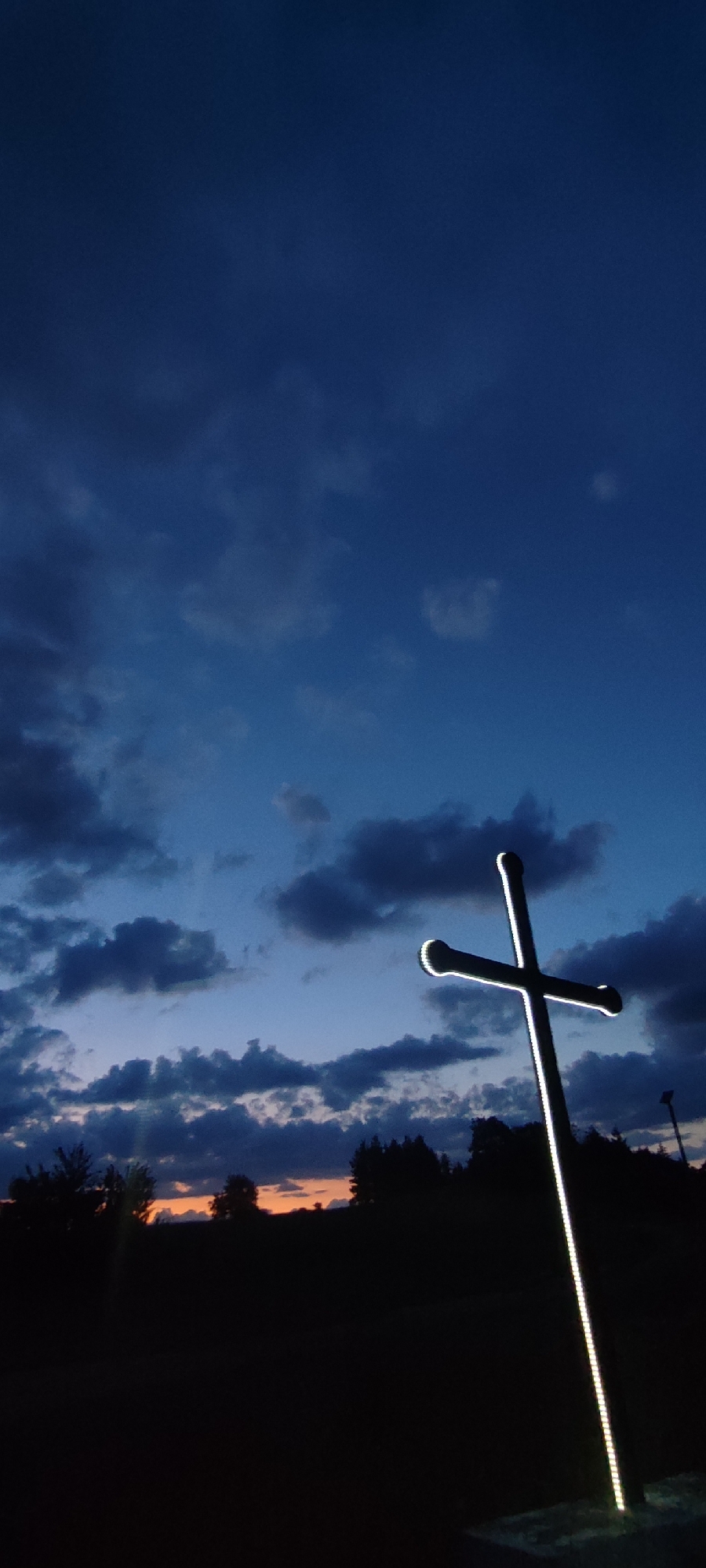 A large, slender Latin cross, illuminated along its edges with a continuous strip of bright white light, stands prominently on the right side of the frame. The cross, which appears dark and silhouetted, extends from the bottom quarter of the image towards the upper right. The sky, occupying the majority of the upper frame, transitions from a deep twilight blue at the top to a lighter blue closer to the horizon. Numerous dark, scattered cumulus clouds are visible across the sky, particularly concentrated in the middle and lower-left sections. A distinct band of orange and yellow light, indicative of a sunset, is visible along the distant horizon, positioned below the cloud cover. This horizon line is defined by a dark, silhouetted expanse of trees and possibly hills. The foreground is entirely in deep shadow, rendering any ground features indistinguishable. A secondary, dark vertical structure, possibly a distant light pole, is faintly visible in silhouette further back on the far right. The scene is set in an outdoor, open environment, likely in or near the city of Gutsal, Bulgaria, under low-light conditions at dusk. No individuals, animals, or specific actions are present.