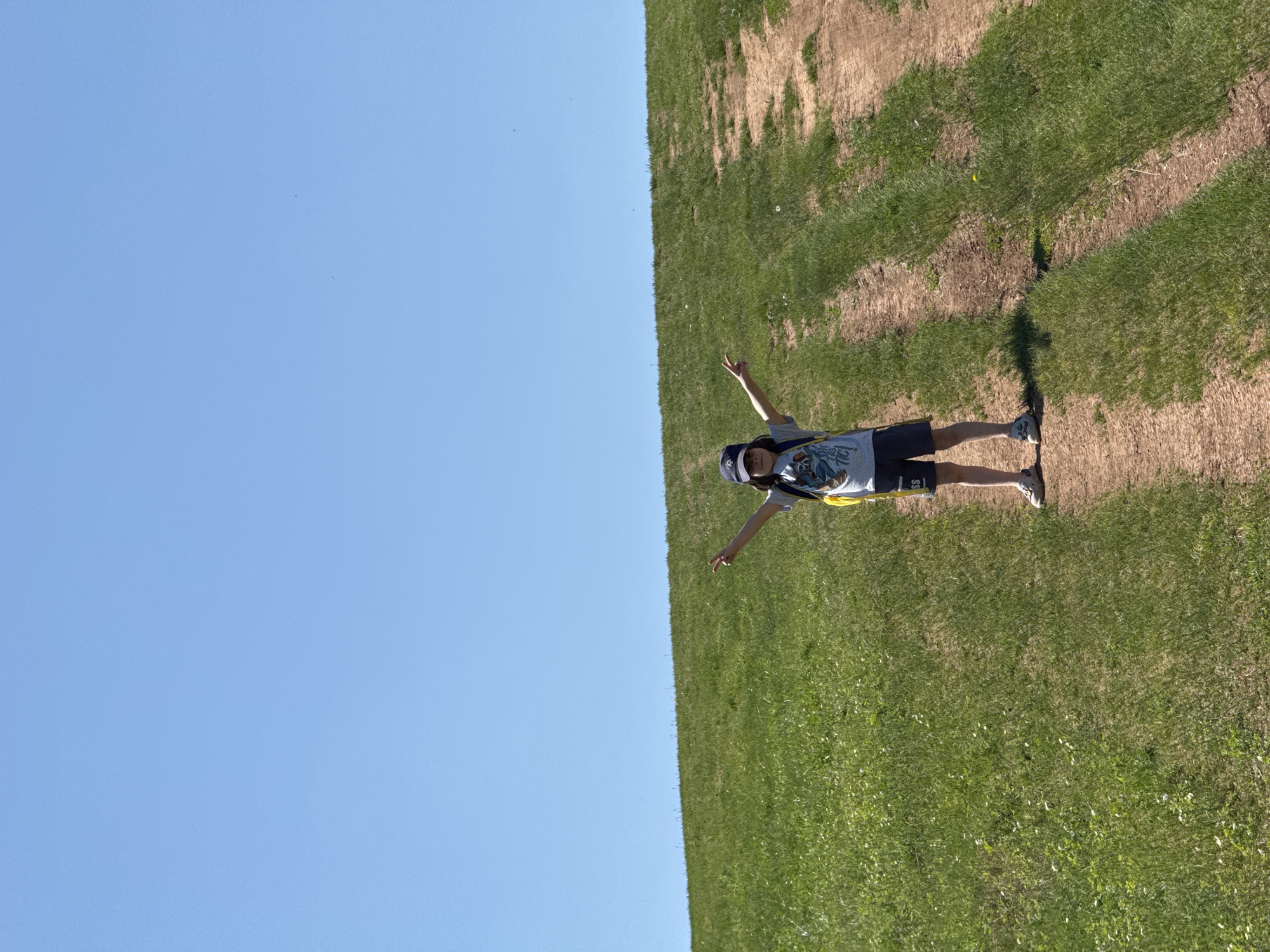 A sunny outdoor scene shows a person standing with their arms outstretched on a grassy slope. The person is wearing a hat, t-shirt, shorts and sneakers. The background consists of a clear blue sky and a green grassy slope with patches of brown earth. The location is not specified in the image.