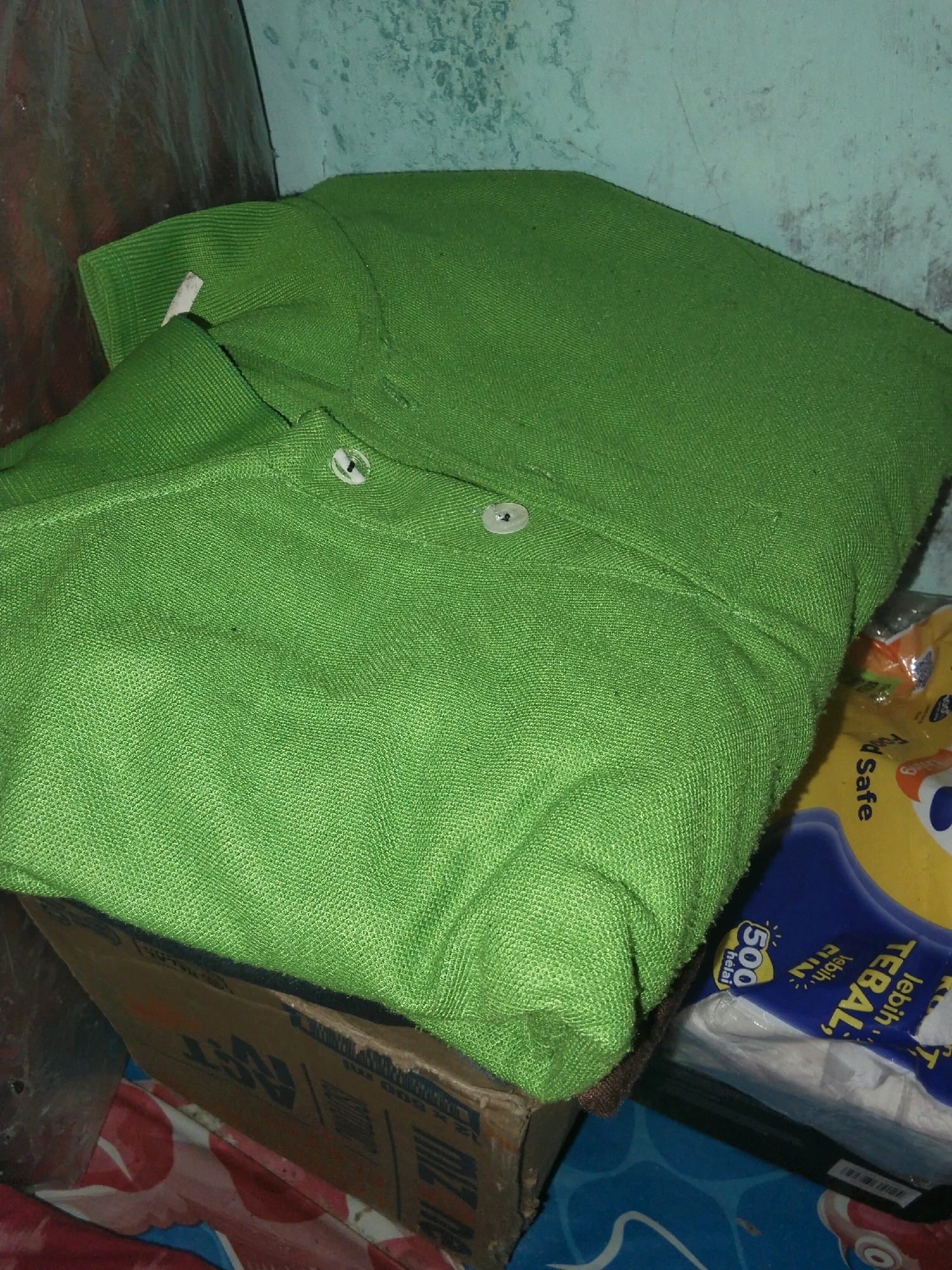 The image shows a folded green polo shirt on top of a cardboard box. To the right of the shirt and box, there is a yellow and blue package of tissues. The background appears to be a textured wall. The scene is dimly lit. The location is identified as Moro, Indonesia.
